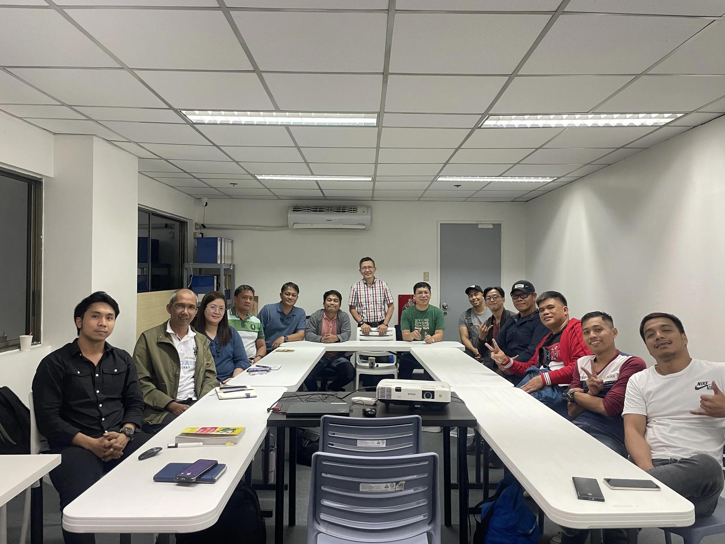 A group of people sitting around a U-shaped conference table in an office setting, with notebooks and a projector on the table. The room has fluorescent lighting and white walls. All individuals are smiling, and they appear to be in a meeting or training session.