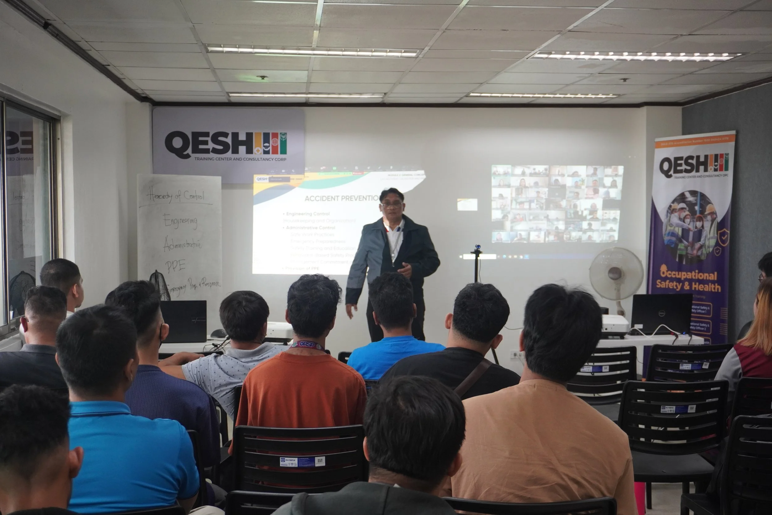 A safety training session in progress with a presenter speaking to an audience. Participants are seated and a projection screen displays information about accident prevention. There is a banner labeled "QESH Occupational Safety & Health" nearby.