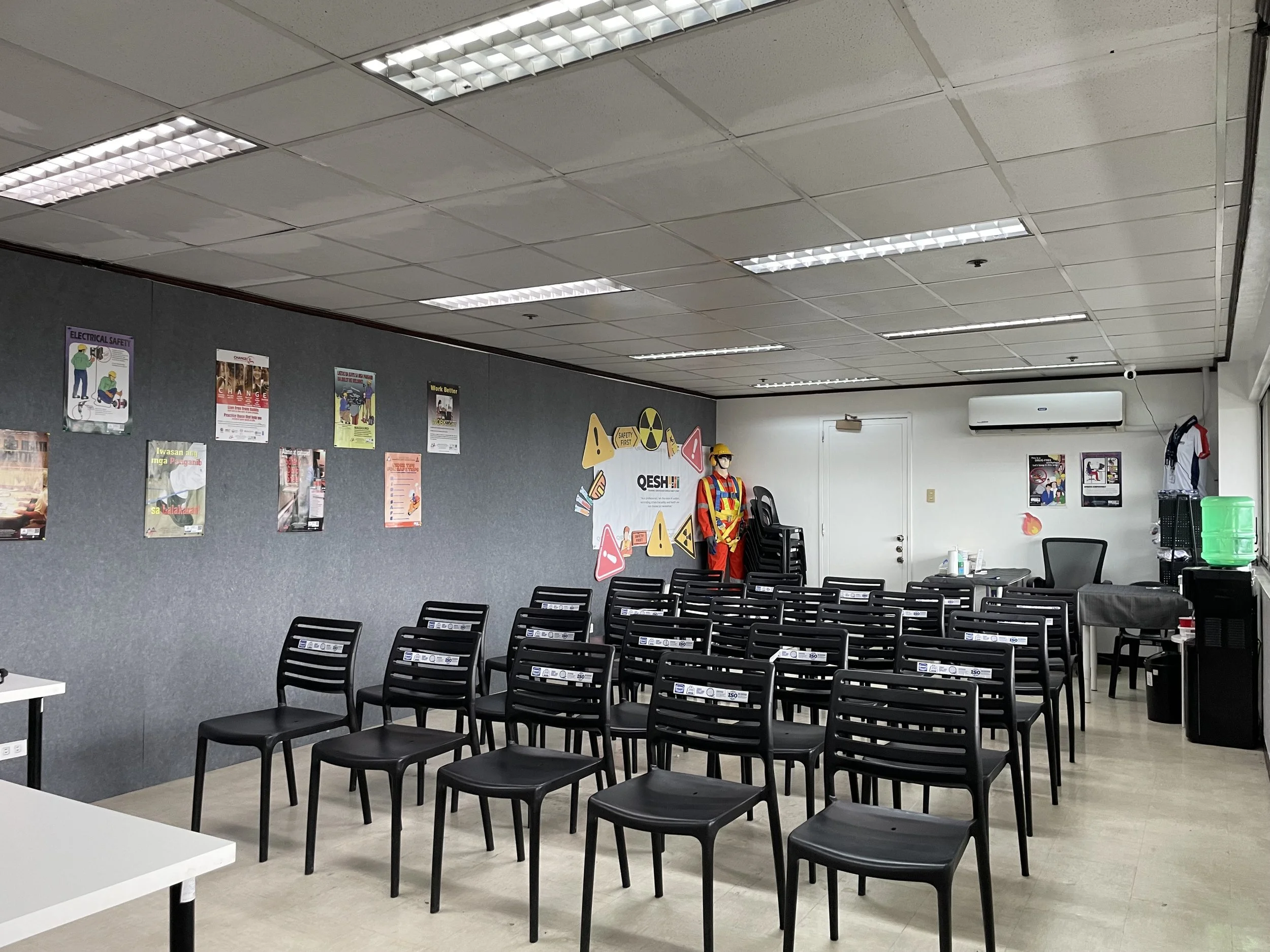 An empty training room with rows of black chairs facing a front wall. The wall features various safety posters and a display with safety gear. A mannequin dressed in safety attire stands in front of the wall. A white table and a desk with chairs are situated at the front. The room is lit with overhead fluorescent lighting.