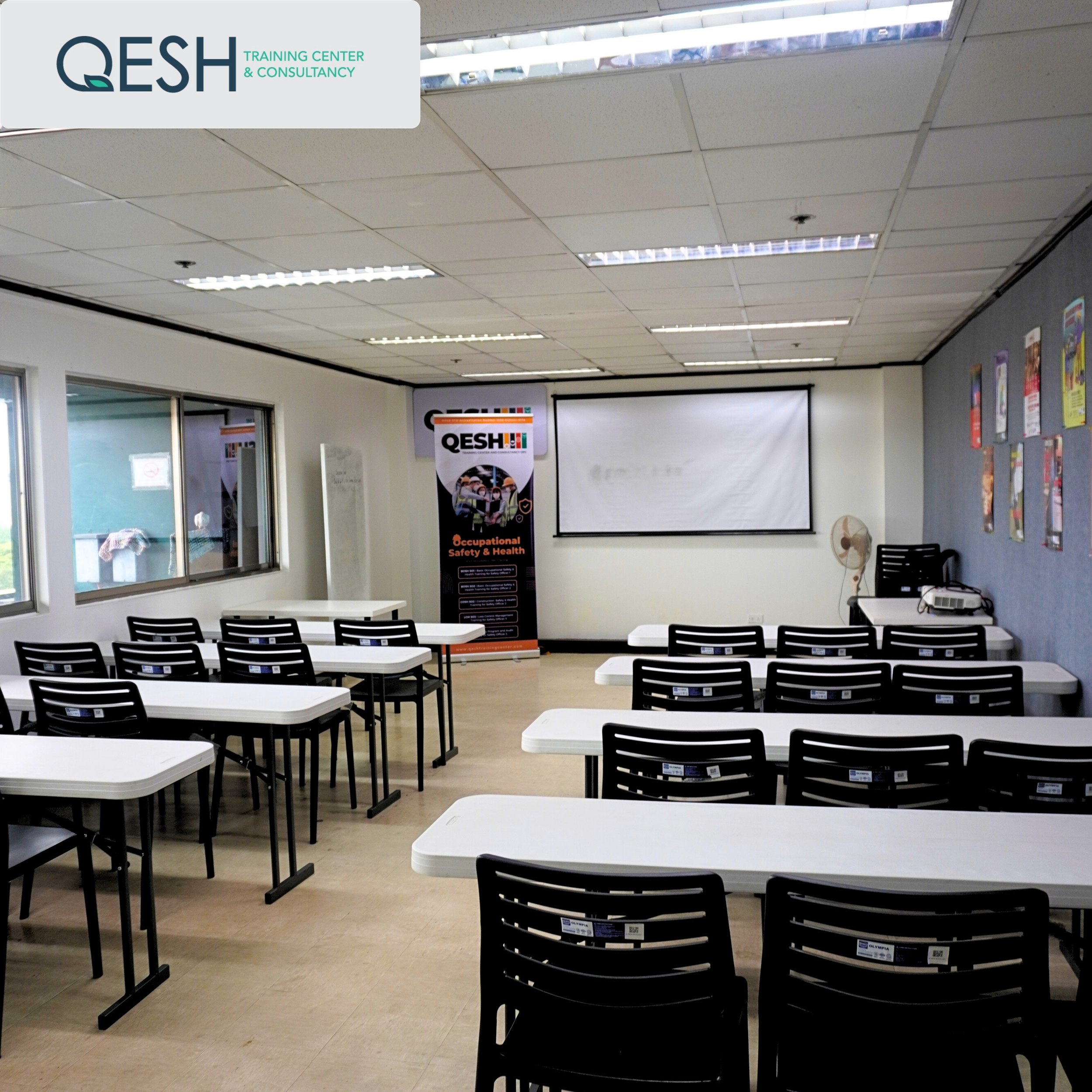 Training room with white desks and black chairs, a projector screen, and posters on the wall.