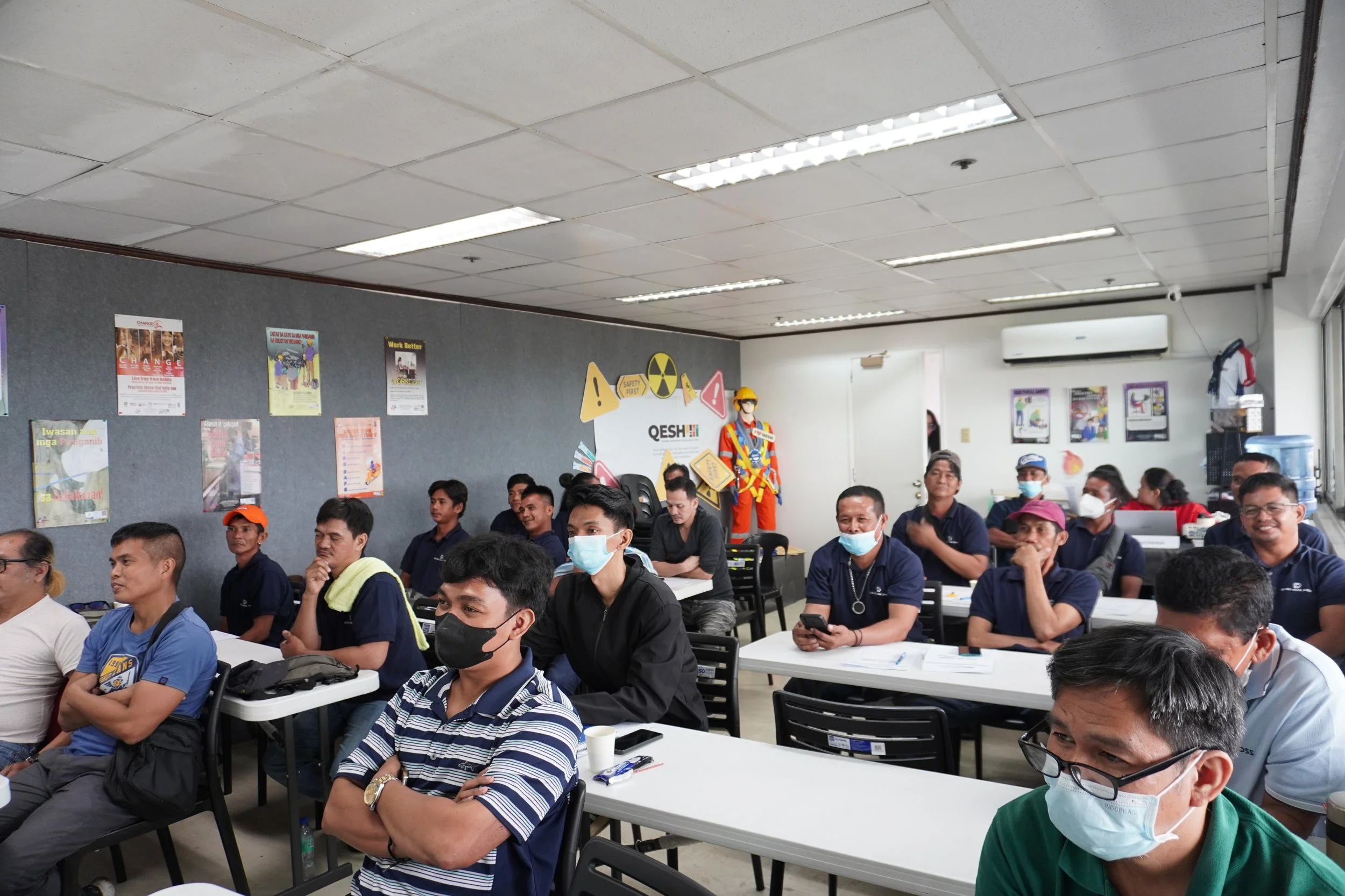 A group of people wearing masks seated in a classroom setting, engaged in a presentation. Posters with safety messages are visible on the walls, and a mannequin dressed in safety gear is in the background.