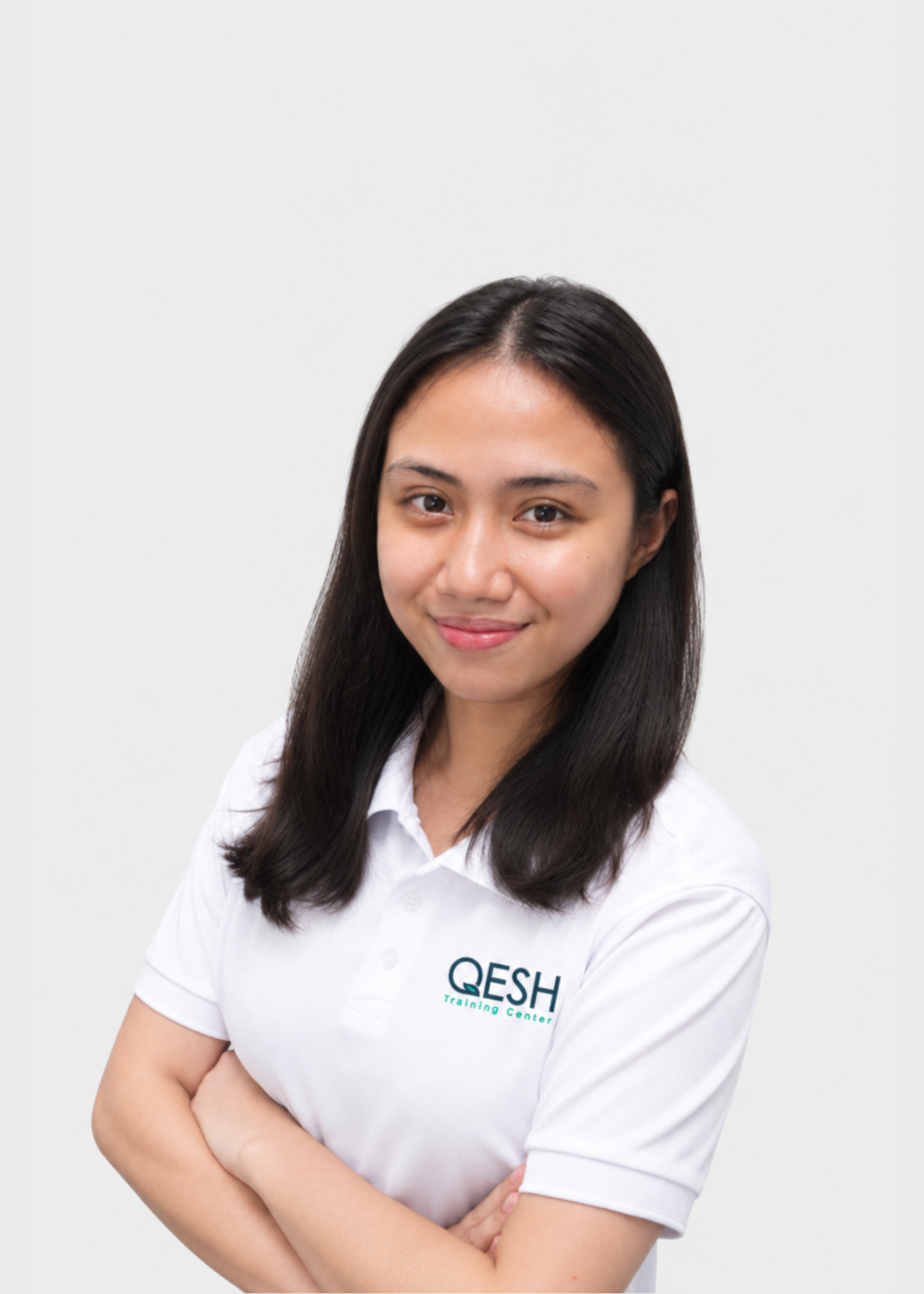 Woman with long black hair, wearing a white polo shirt and a blue lanyard, smiling with arms crossed in front of a plain background.