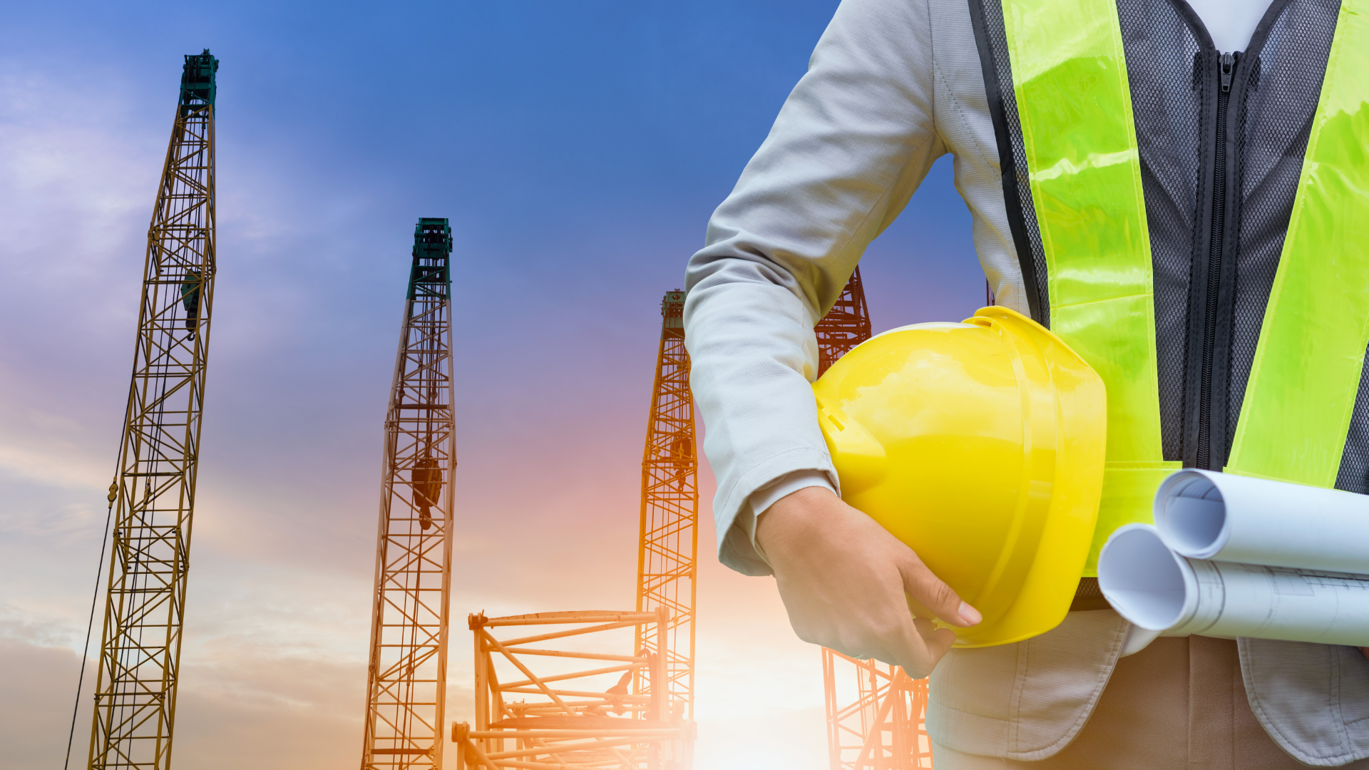 Construction worker holding yellow hard hat and blueprints at a construction site with cranes and scaffolding during sunset.