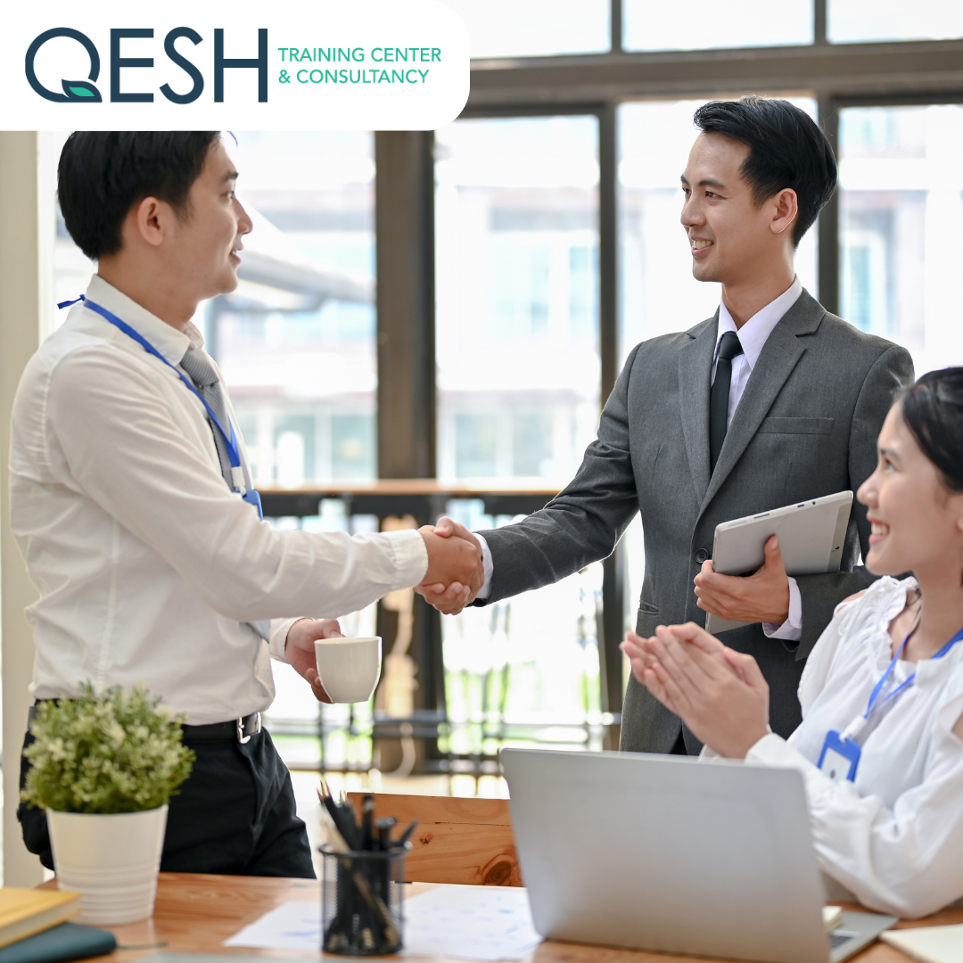 Three business professionals in an office setting; two men shaking hands while a woman sits at a desk with a laptop, smiling and clapping. A potted plant and office supplies are visible on the desk. The logo "QESH Training Center & Consultancy" is in the upper left corner.