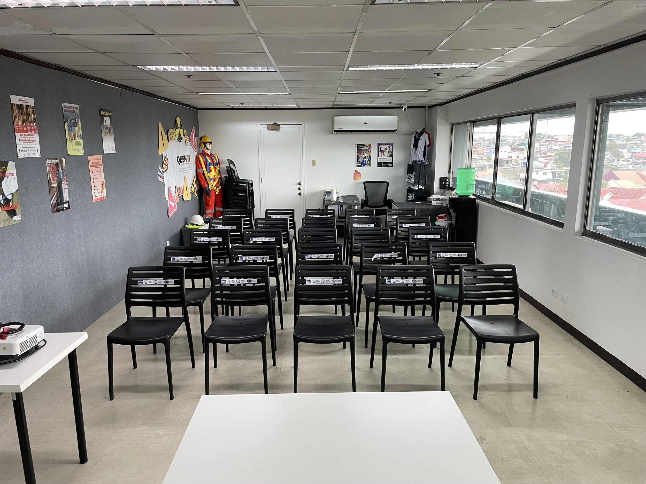 Empty classroom with rows of black chairs, a mannequin wearing safety gear in the corner, posters on the wall, and windows showing a cityscape.