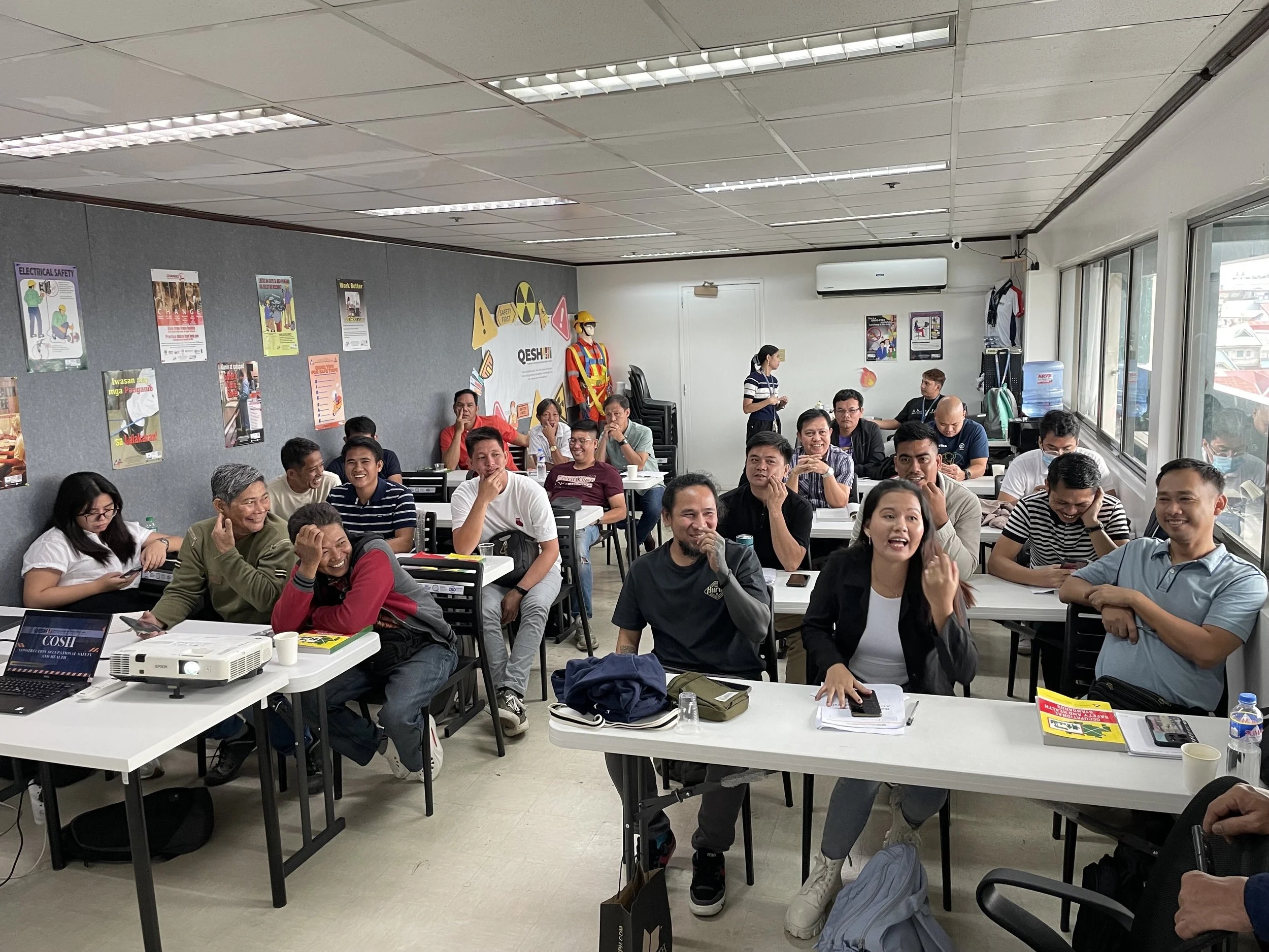 A classroom filled with adults sitting at desks, engaged in a training session. The room is well-lit with posters on safety and health hung on the walls. A projector is on a desk at the front, and the participants appear attentive and interactive.