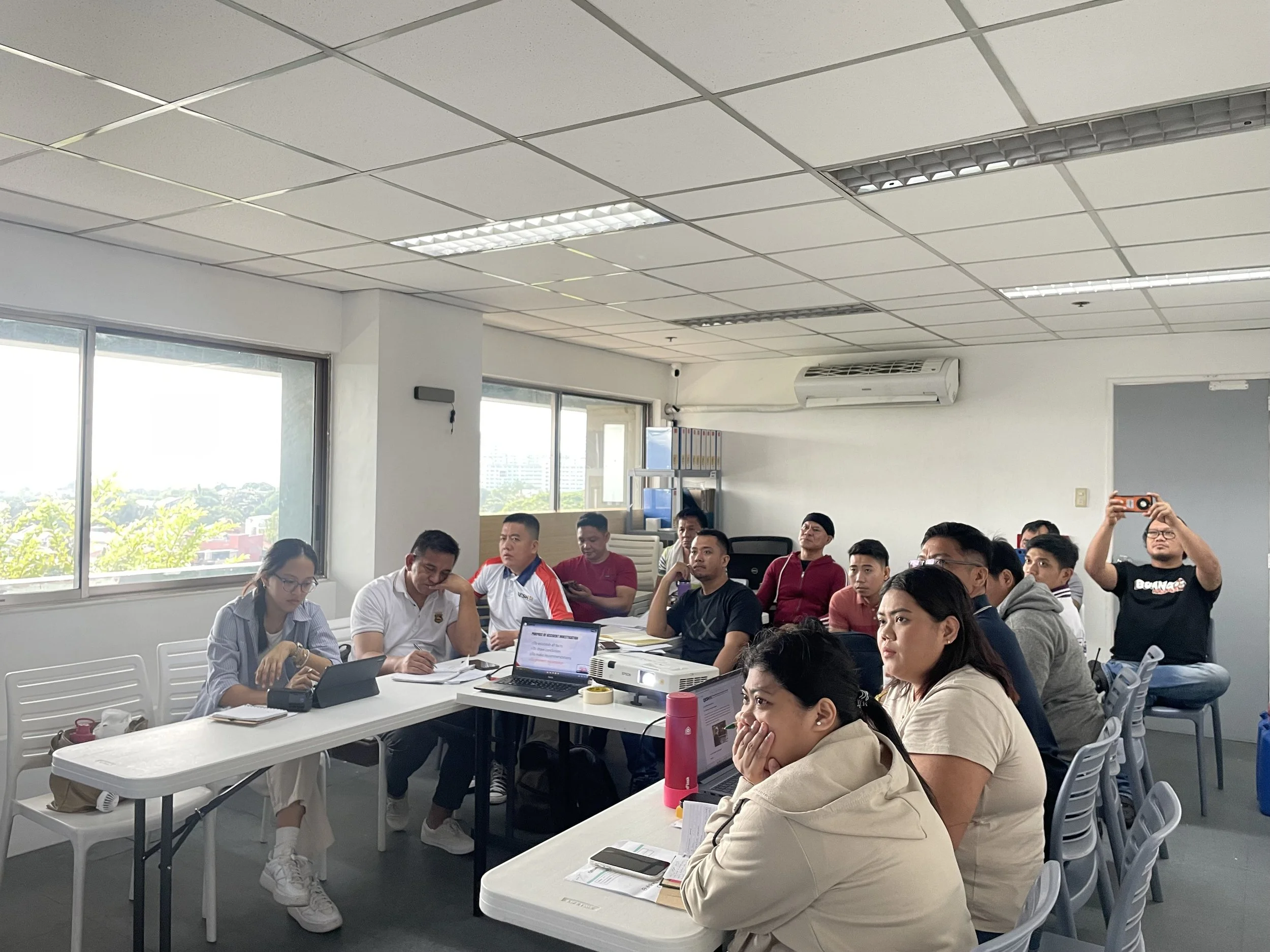 A group of people in a casual classroom setting, sitting around tables with laptops, notebooks, and phones. Some individuals are paying attention to something outside the frame, while one person is using a phone to take a photo. The room has windows with a view outside and is equipped with fluorescent lights and air conditioning.