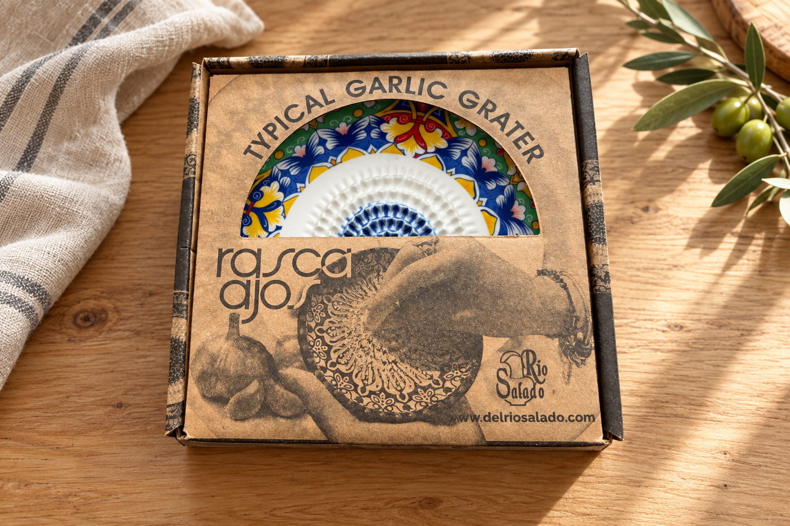 Boxed Spanish garlic grater plate on an oak table with linen and an olive branch