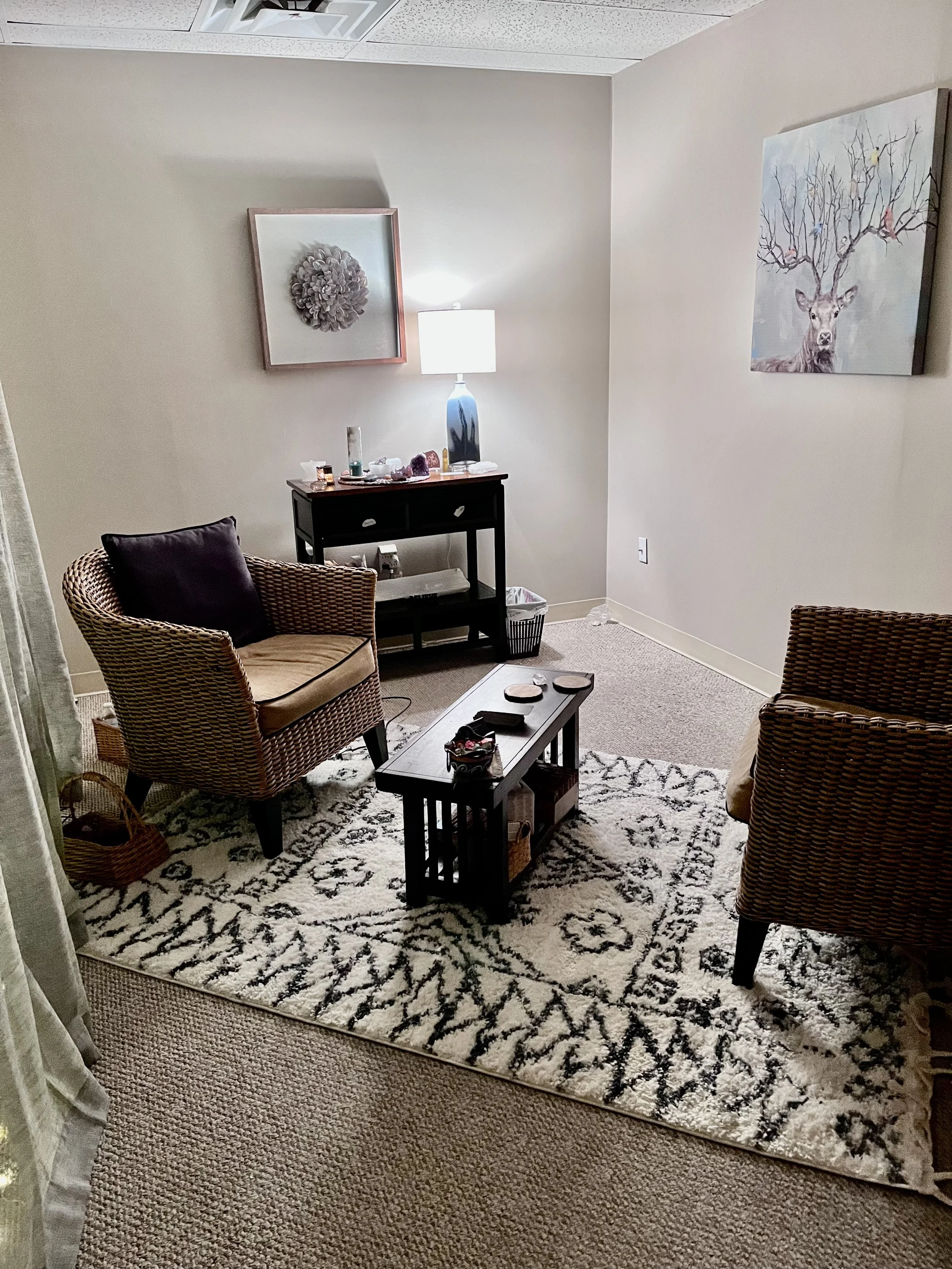 A small living room with two wicker armchairs, a black coffee table, and a black side table. The floor has a patterned rug, and the walls feature artistic decorations, including a deer painting and a flower artwork. There is a lamp on the side table, and various small objects are on the tables.