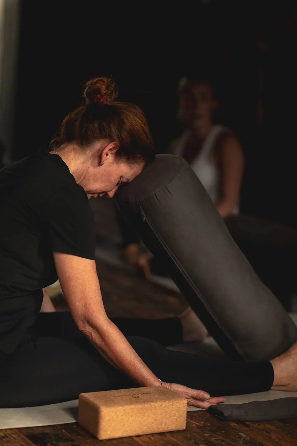 Woman practicing yoga on a mat with a yoga block and bolster in a dimly lit studio.
