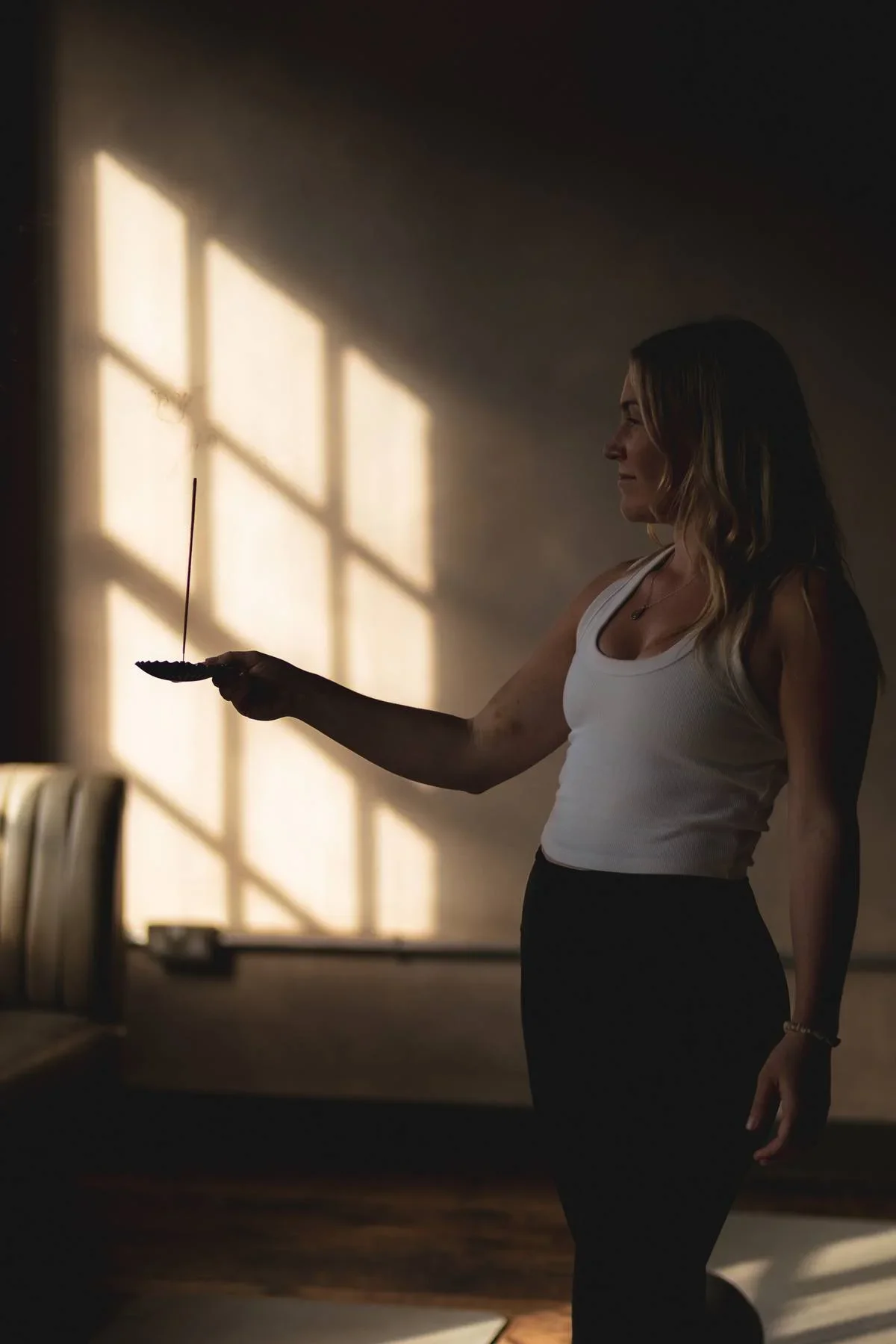 A woman in a white tank top and black pants is holding a feather and an incense stick while kneeling on a yoga mat in a dimly lit room with sunlight streaming through a window.