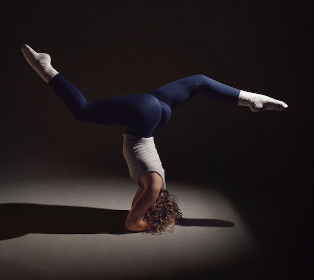 A woman performing a headstand yoga pose in a dark room, with her body balanced upside down and legs extended horizontally.