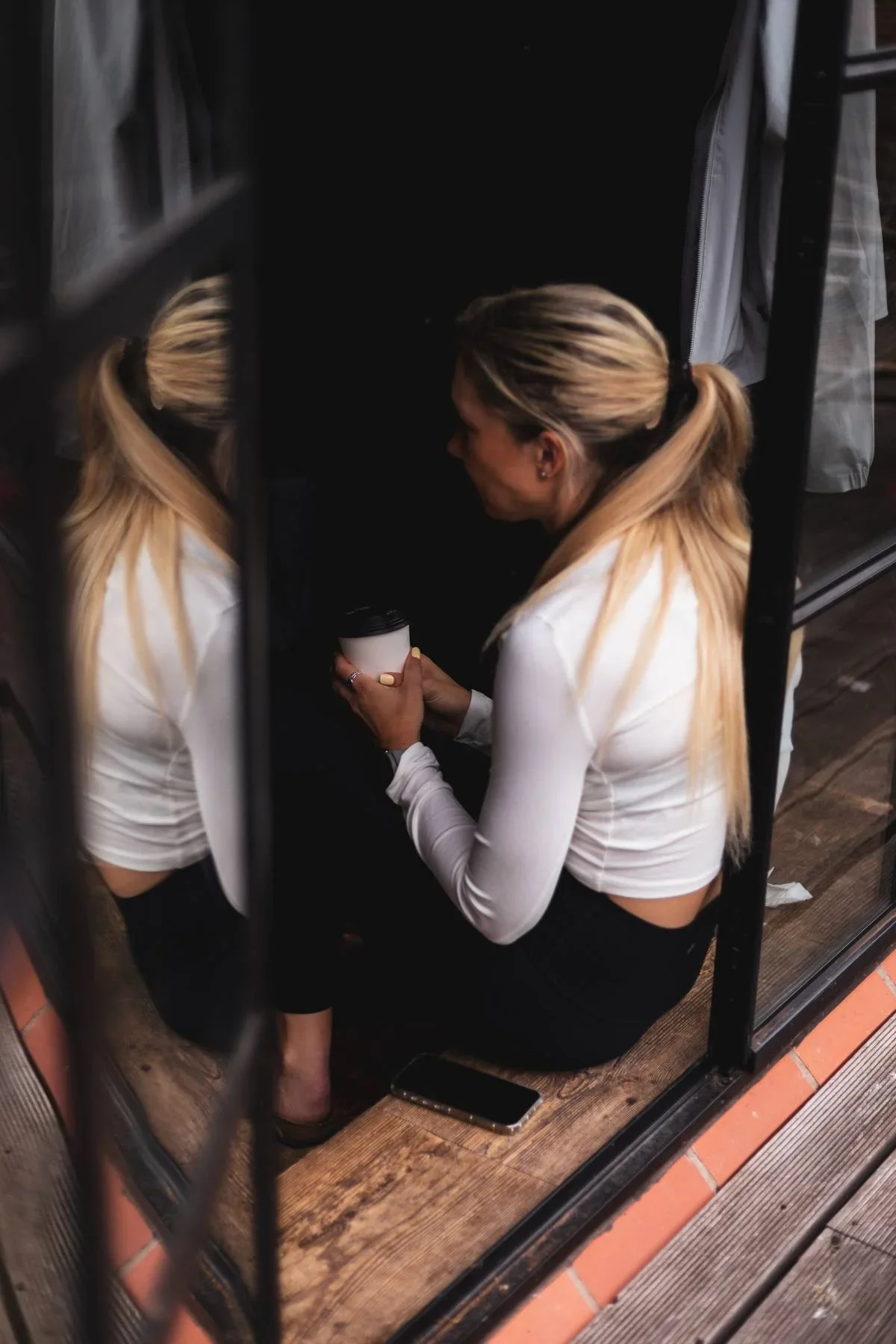 A woman with long blonde hair tied in a ponytail, wearing a white long-sleeve top and black pants, sitting on a wooden floor inside a small enclosed space, holding a white disposable coffee cup, viewed through a window or glass panel with metal frami
