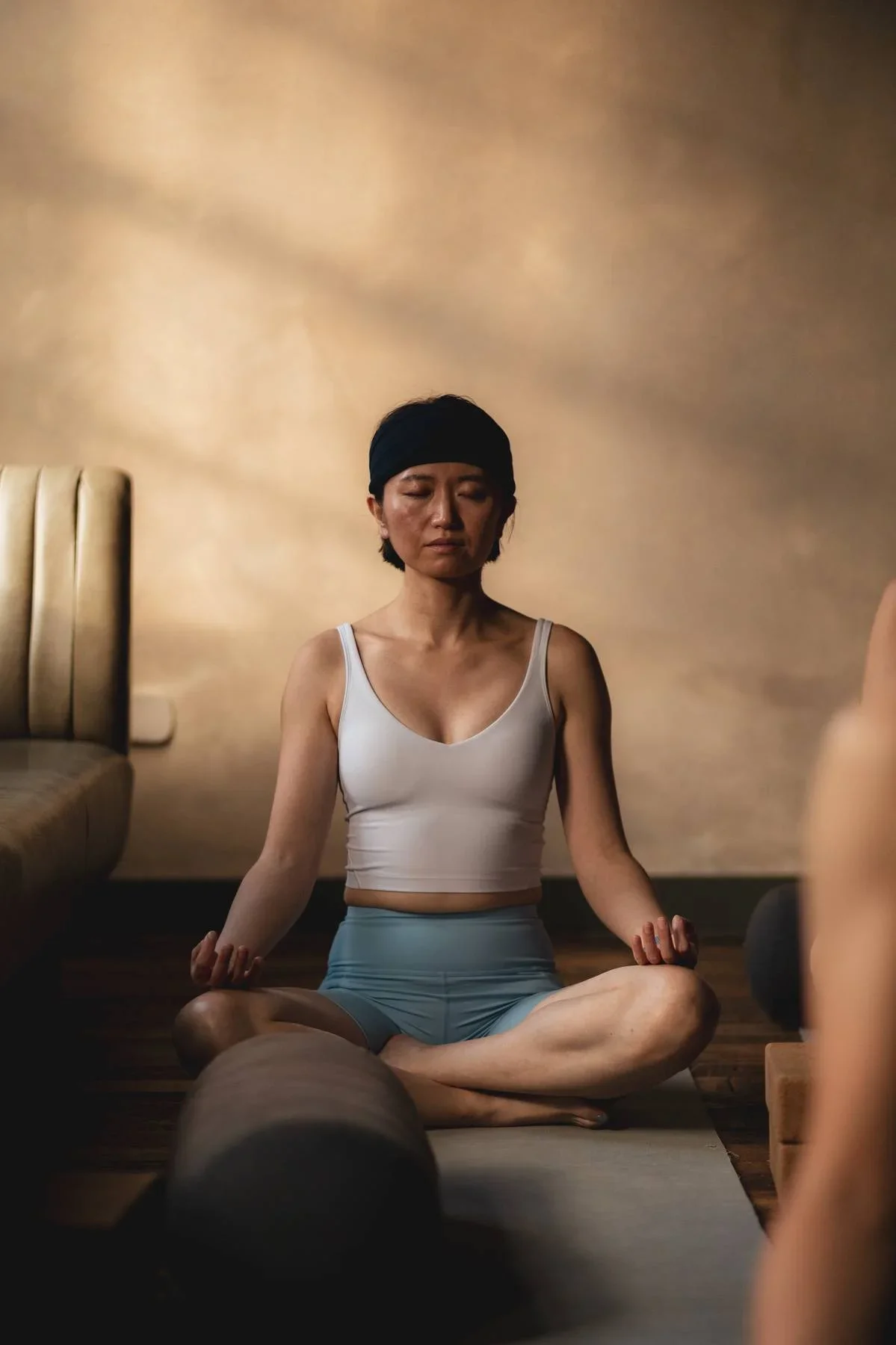 A woman meditating in a cross-legged position, sitting on a yoga mat in a softly lit room.