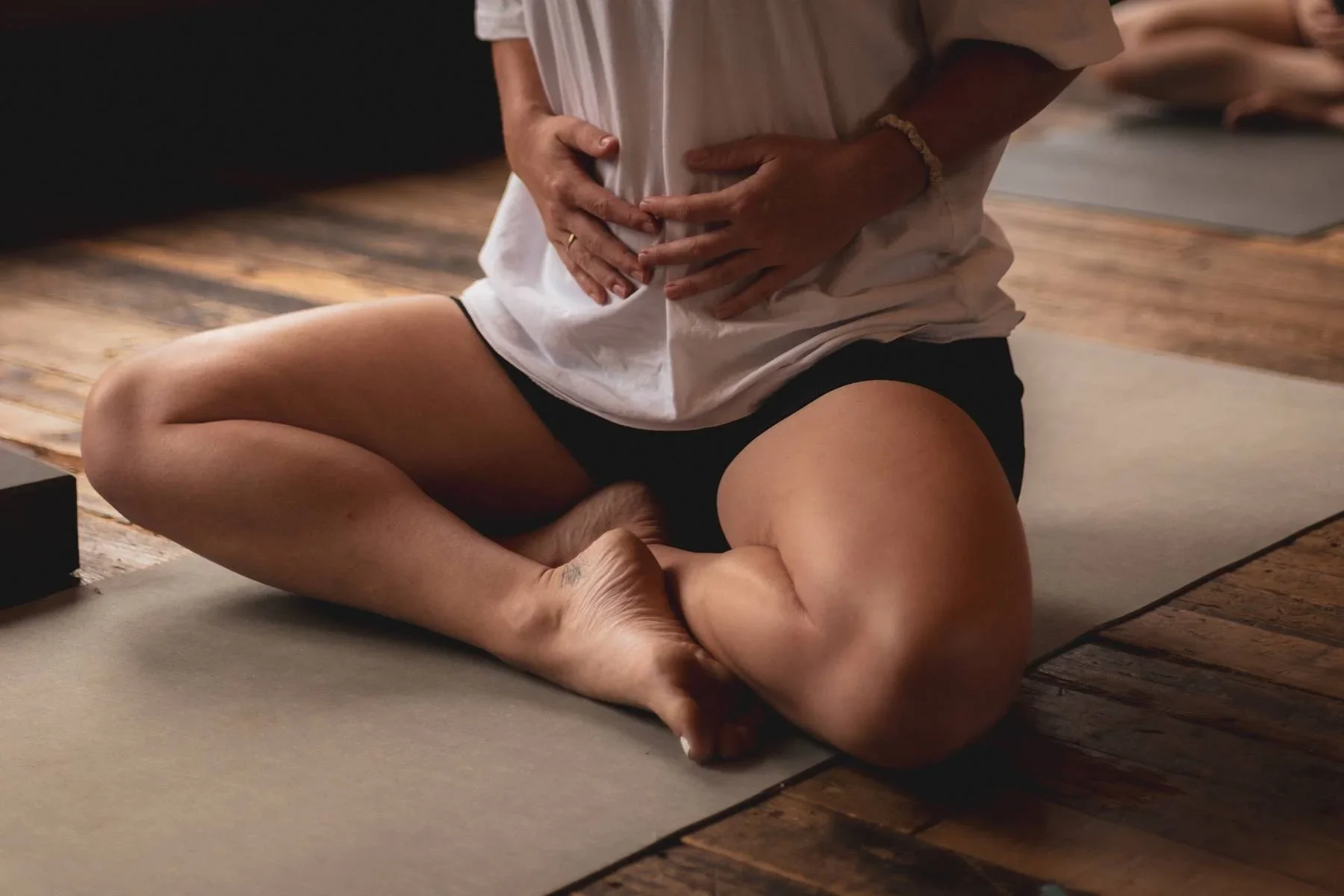 Yoga practitioner seated cross-legged on a mat with hands resting on the abdomen, practising mindful breathing in a calm studio setting.