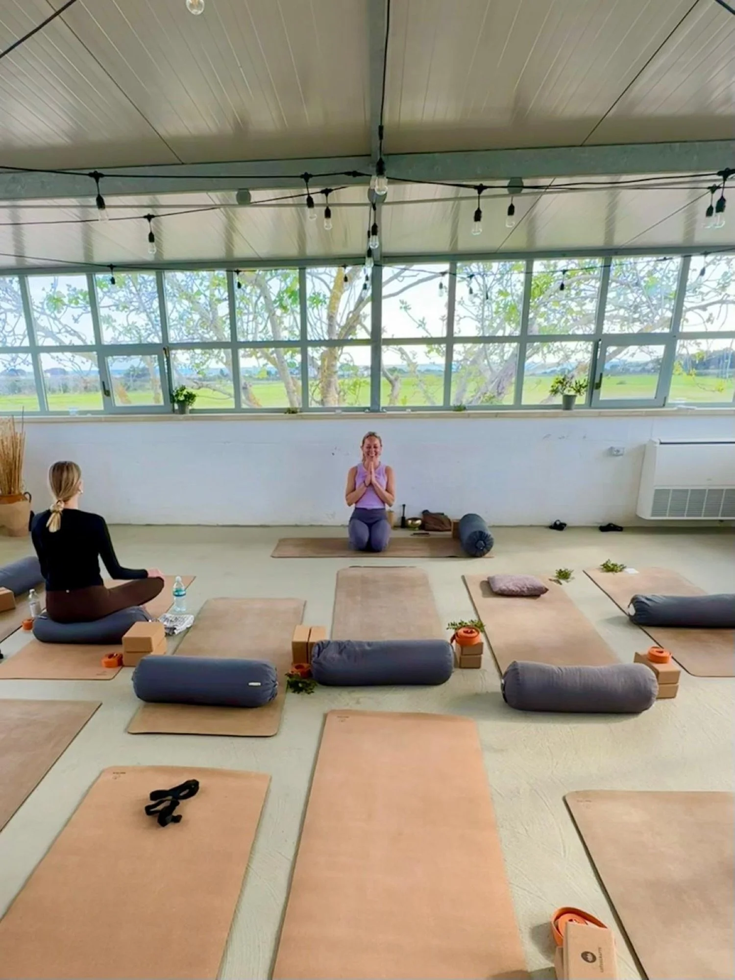 Morning yoga practice at Masseria Specula, a teacher seated at the front of a bright room with mats and bolsters laid out and tall windows onto the Puglian countryside