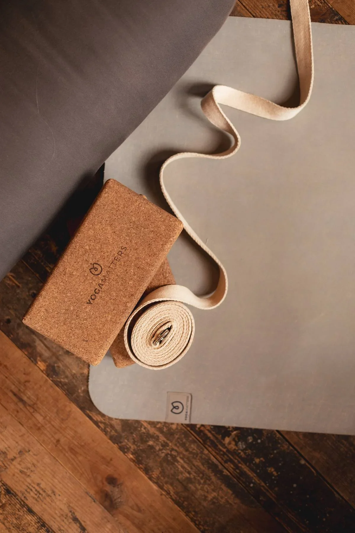 A cork yoga block, a rolled-up beige yoga strap, and a gray yoga mat on a wooden floor.