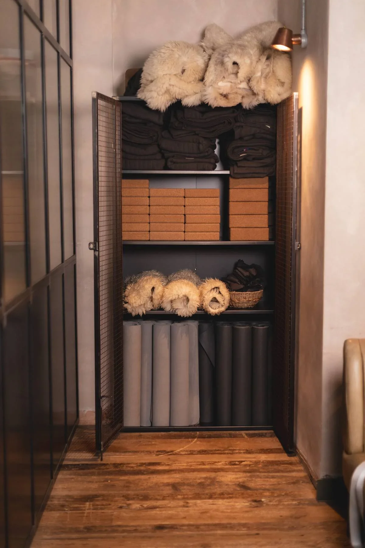 A black shelving unit with multiple shelves, featuring fur hats or scarves, folded brown clothes, stacks of orange and gray foam tiles, and a small woven basket containing dark items, situated against a wall with wooden flooring.