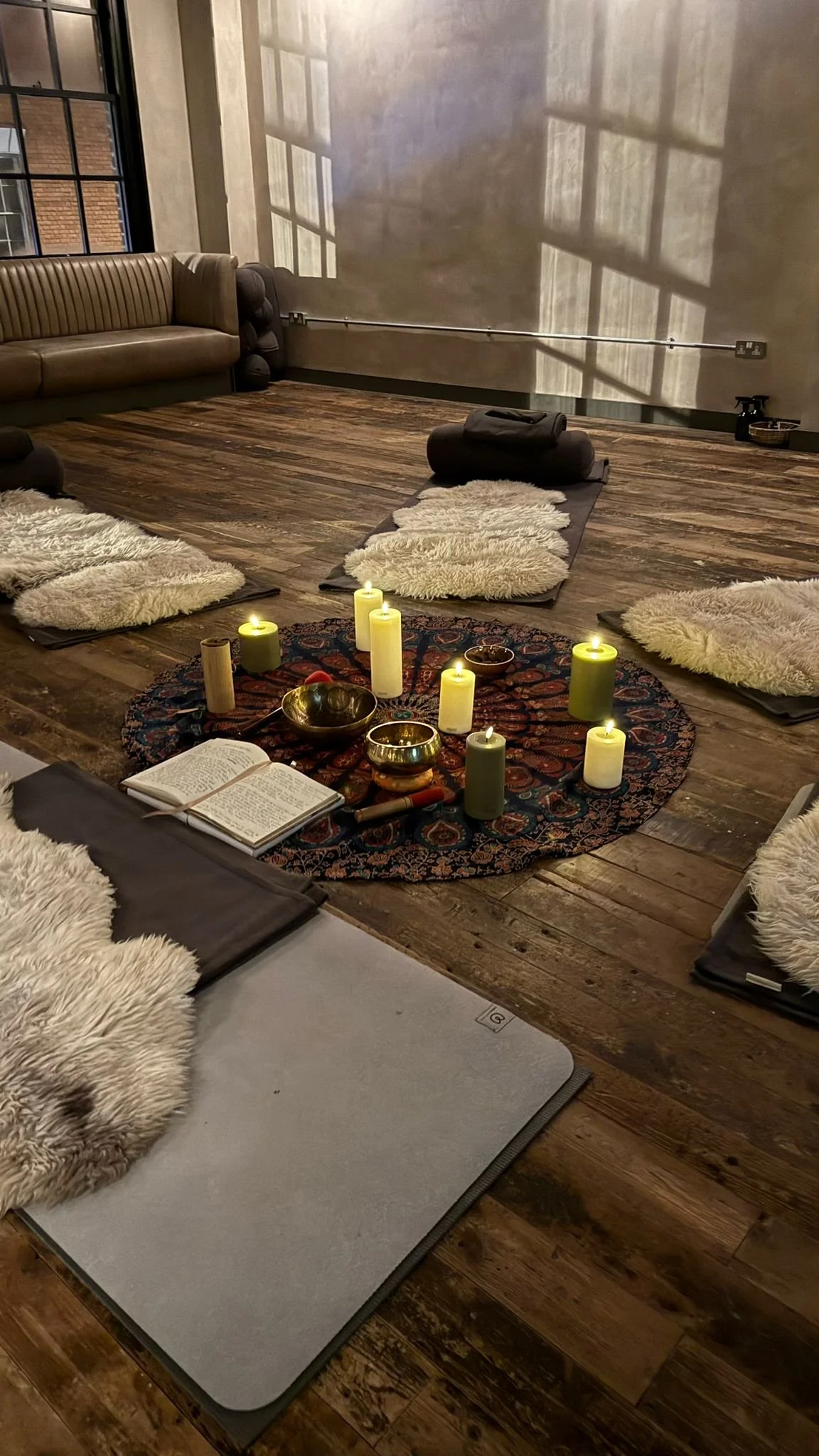 Yoga mats with sheepskin covers, candles, and singing bowls arranged in a circle on a patterned rug in a cozy, loft-style room with wooden floors and large windows
