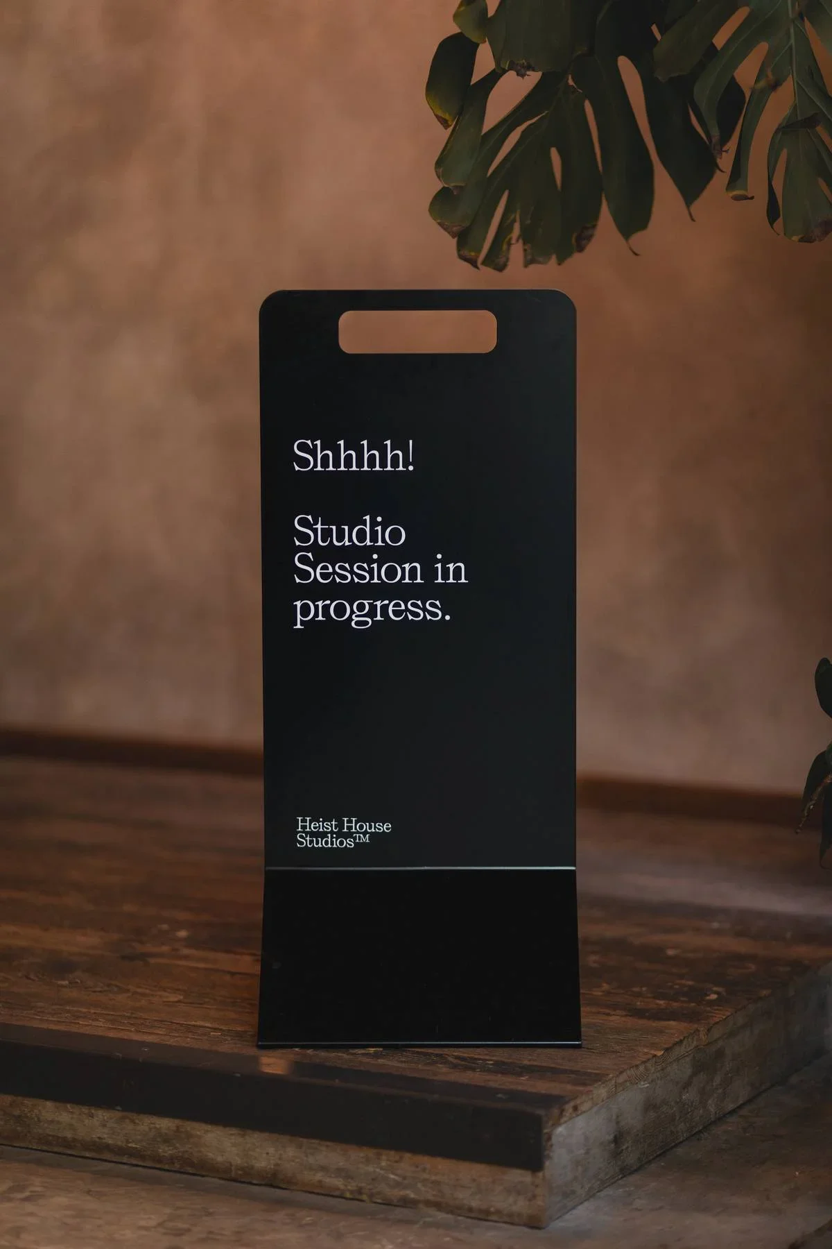 A black sign with white text stating, 'Shhhh! Studio Session in progress. Heist House Studios™', placed on a wooden table with greenery in the background.