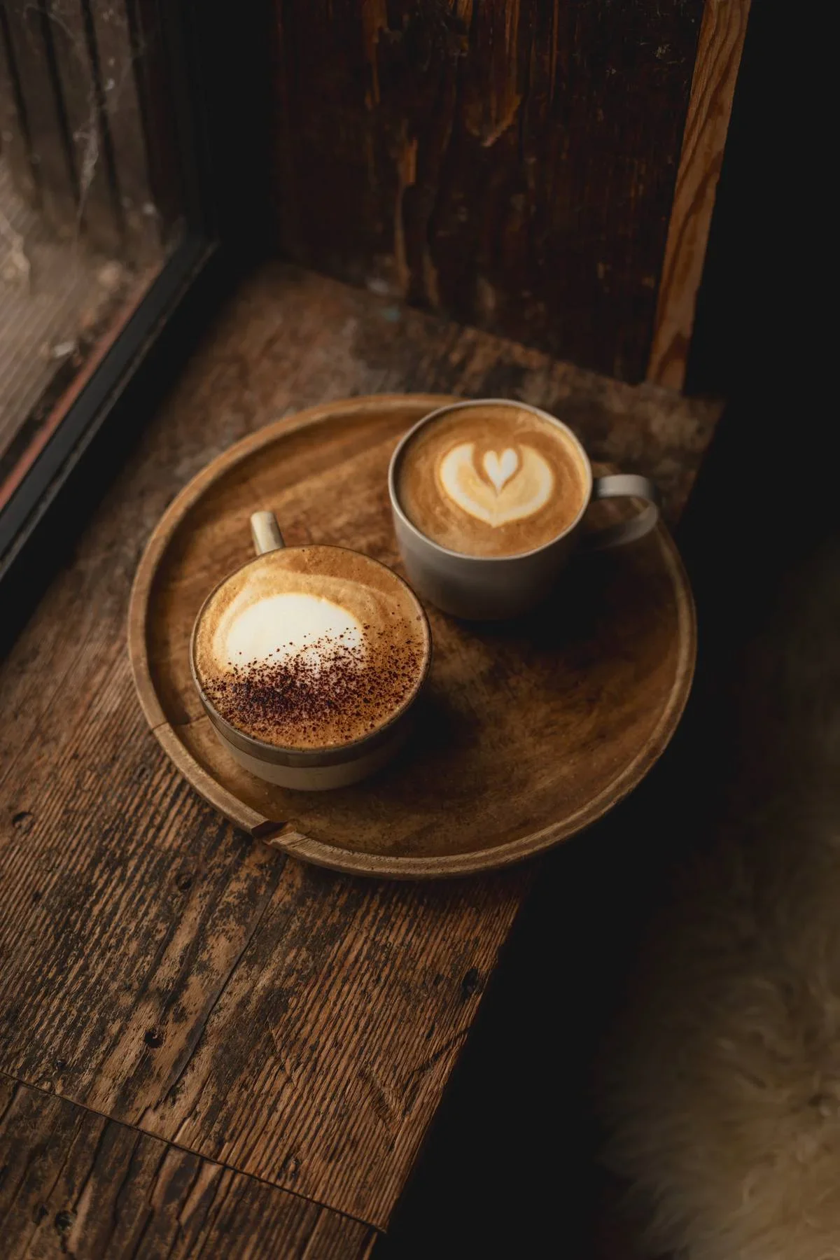 Two cups of coffee with latte art, one with a heart shape and the other with a heart and tulip design, on a wooden tray near a window.