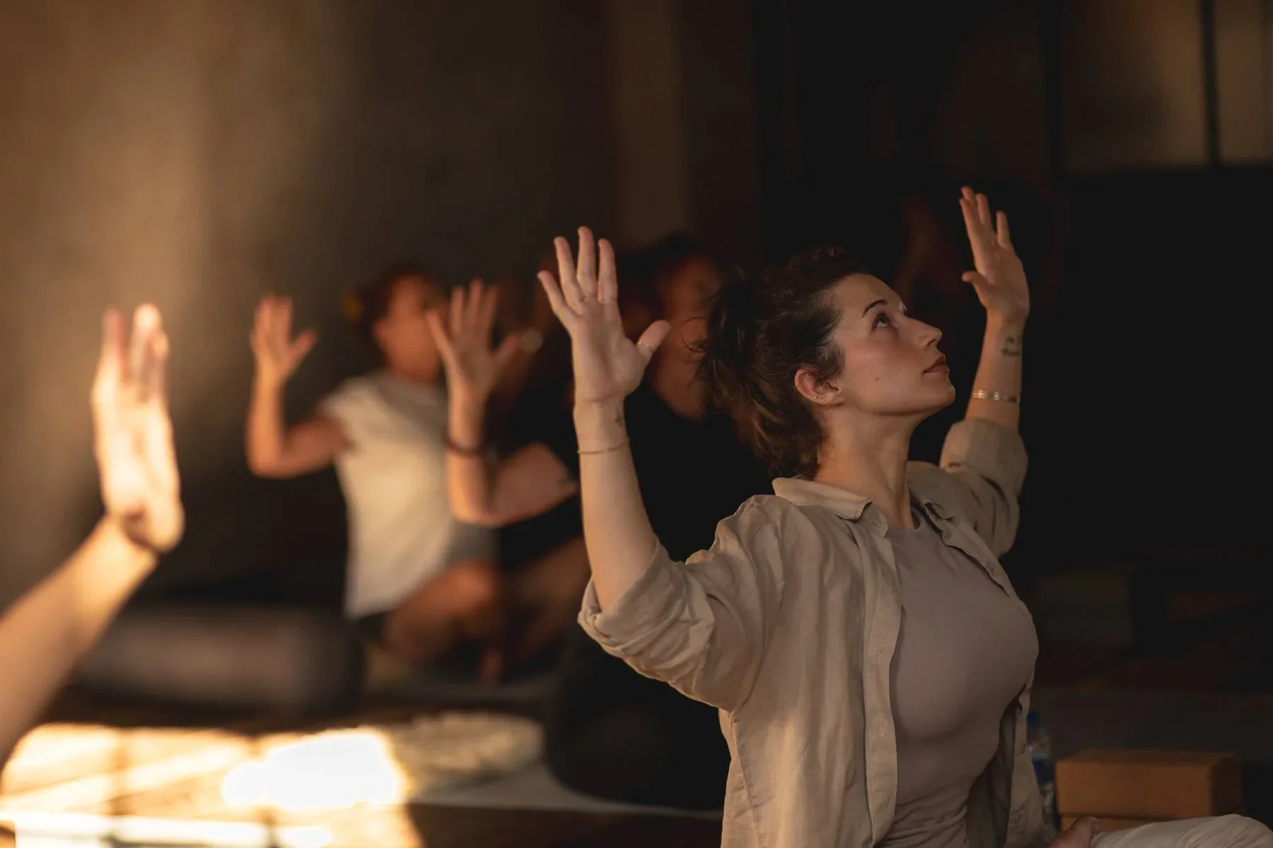 A group of women practicing yoga or meditation, sitting on mats with their eyes closed and hands raised in a mudra, in a dimly lit room.