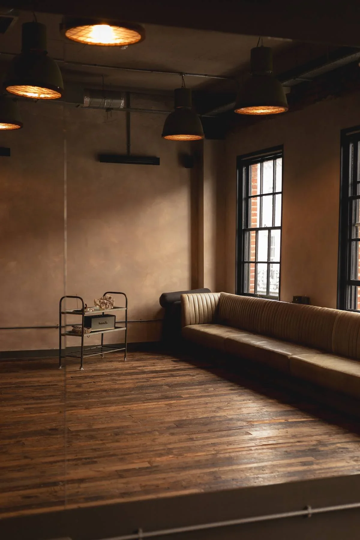 Interior of a dimly lit room with wooden flooring, a long beige leather couch, and large windows with black frames. There are five black pendant lights hanging from the ceiling.