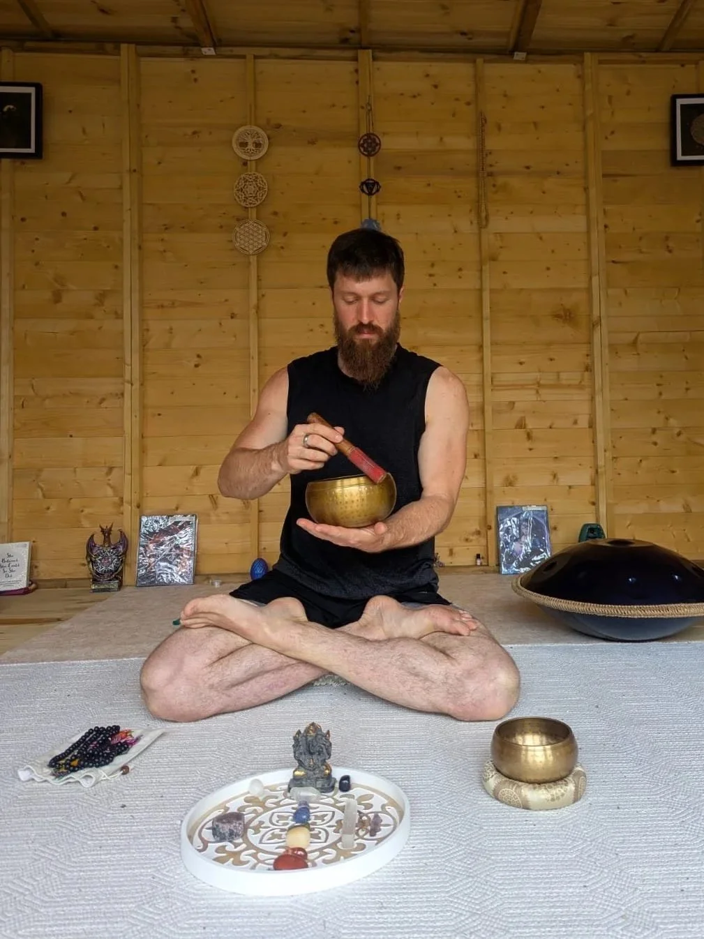 A man with a beard sitting cross-legged on a mat in a wooden room, playing a singing bowl with a mallet. There are various objects including another bowl, jewelry, and decorative items, with framed pictures hanging on the wooden wall behind him.