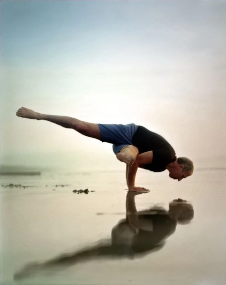Larry Schultz, the founder of Rocket yoga, holding a crow pose arm balance on a reflective water surface
