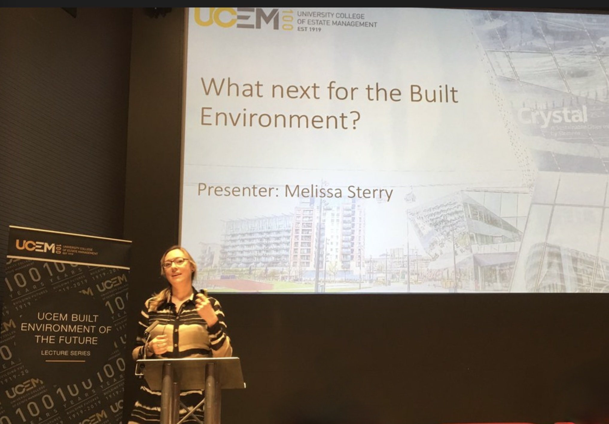 Design scientist Dr. Melissa Sterry giving a presentation at the University College of Estate Management event. The screen displays the title 'What next for the Built Environment?' with the presenter's name. A banner with UCEM branding is visible.