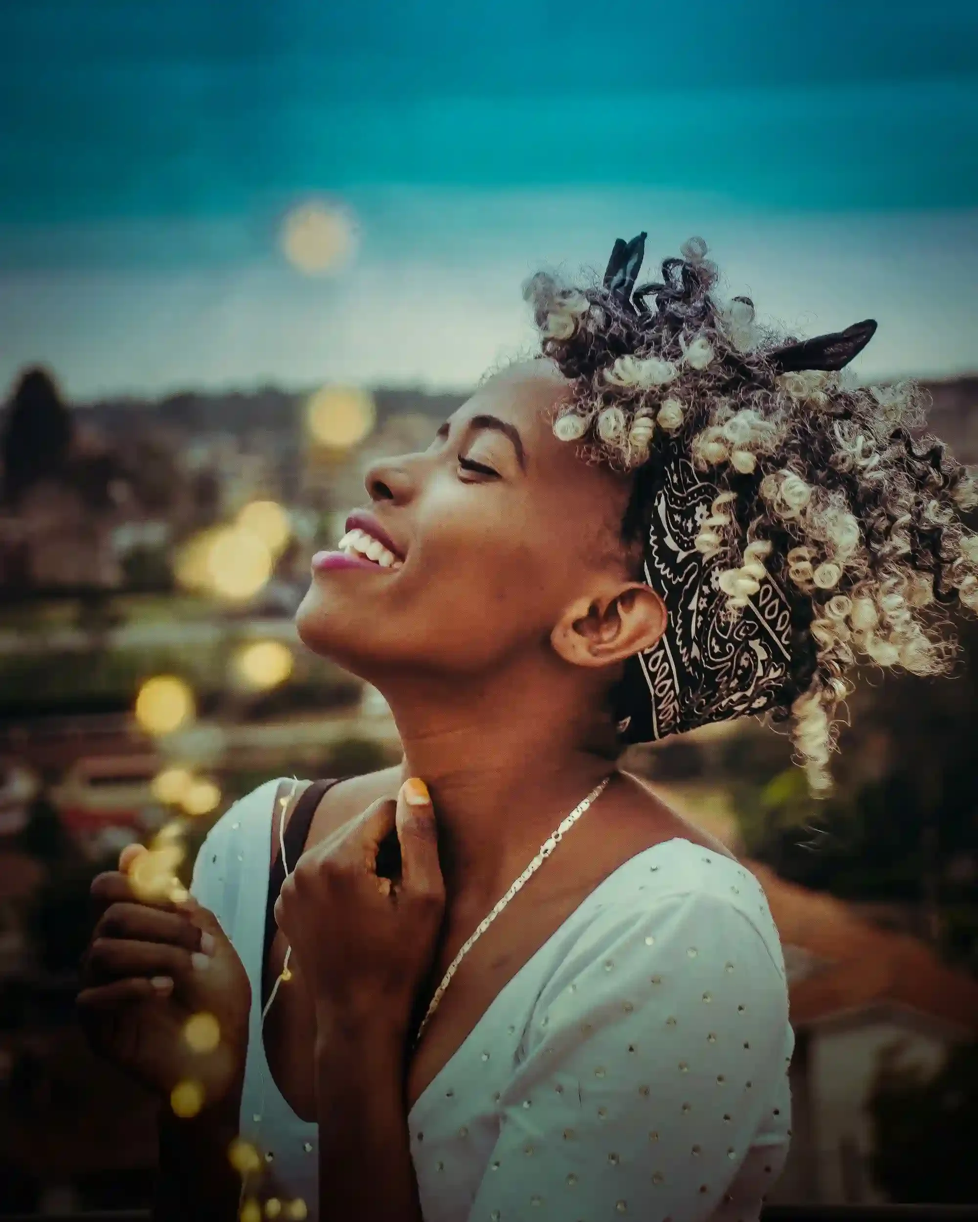 A woman with curly hair styled with shells and small decorative objects, wearing a bandana and a white dress with small dots, is smiling outdoors during the evening with a blurred cityscape and bokeh lights in the background.