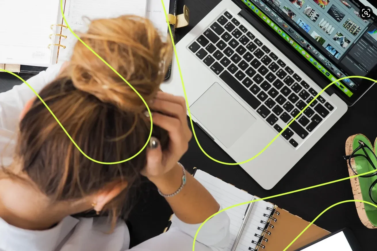 A woman with red hair working at a cluttered desk, looking at a laptop with a video editing software open, surrounded by notebooks, glasses, and a green tray.