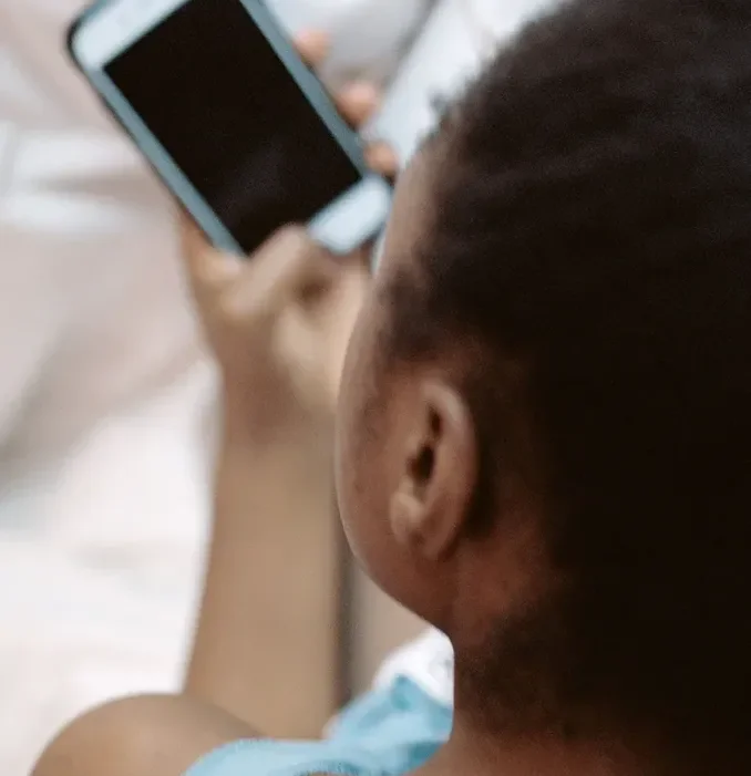 A child with short hair sitting on a bed, holding and looking at a smartphone.