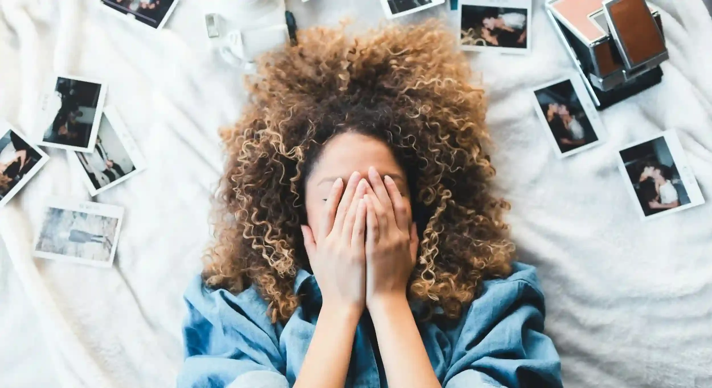 A woman lying on a white surface surrounded by scattered instant photographs, covering her face with her hands, with curly hair and wearing a blue top.