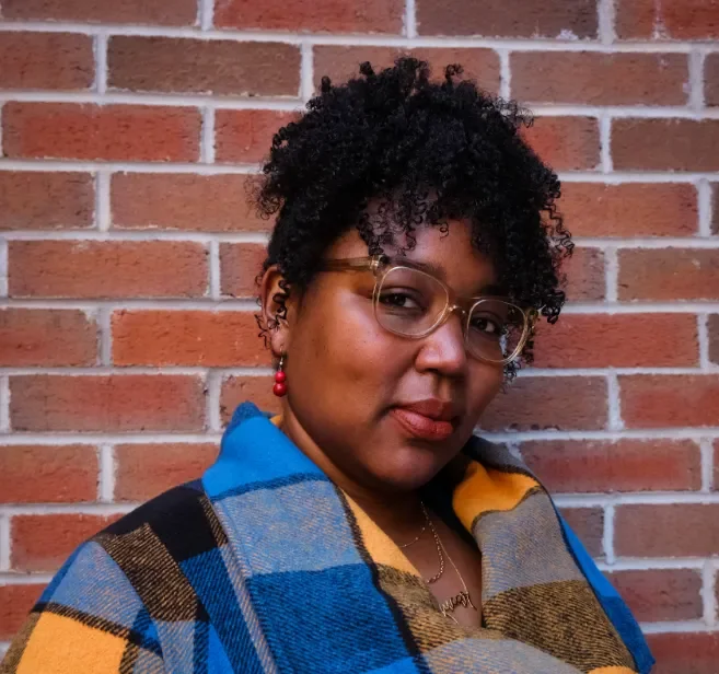 A woman with short curly black hair wearing glasses, red earrings, and a colorful plaid coat standing against a brick wall.