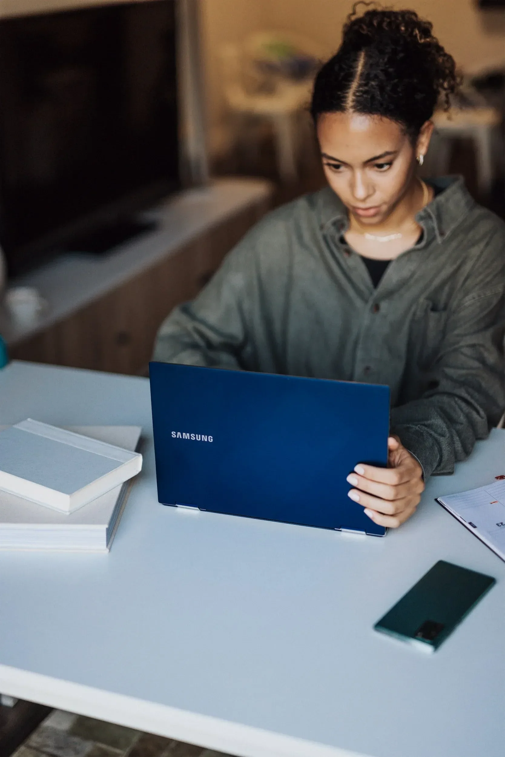 A woman with curly hair and earrings sitting at a table, examining a blue Samsung booklet, with a smartphone and white boxes on the table.