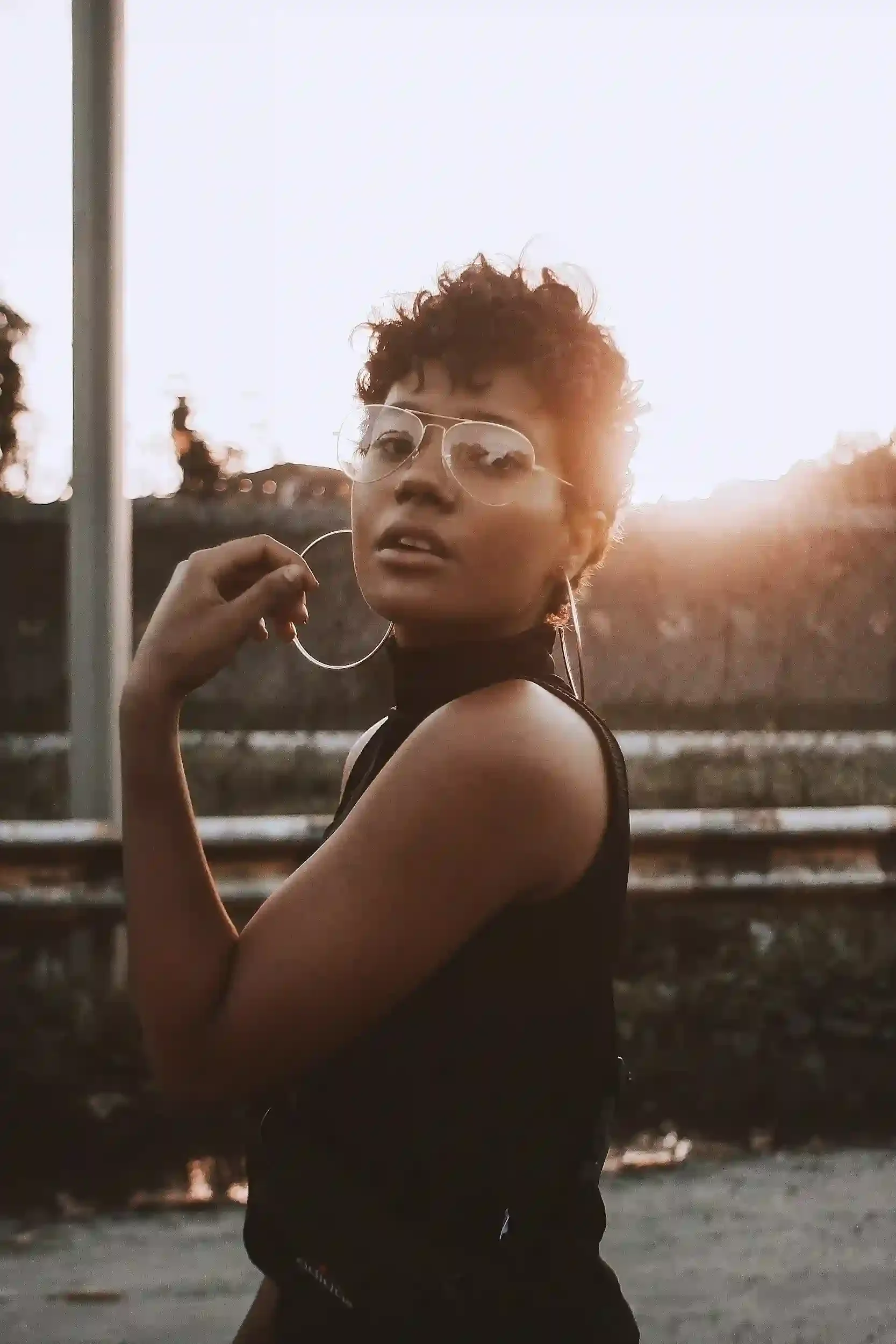A young woman with curly hair wearing aviator sunglasses and hoop earrings, standing outdoors during sunset, near a body of water with trees in the background.