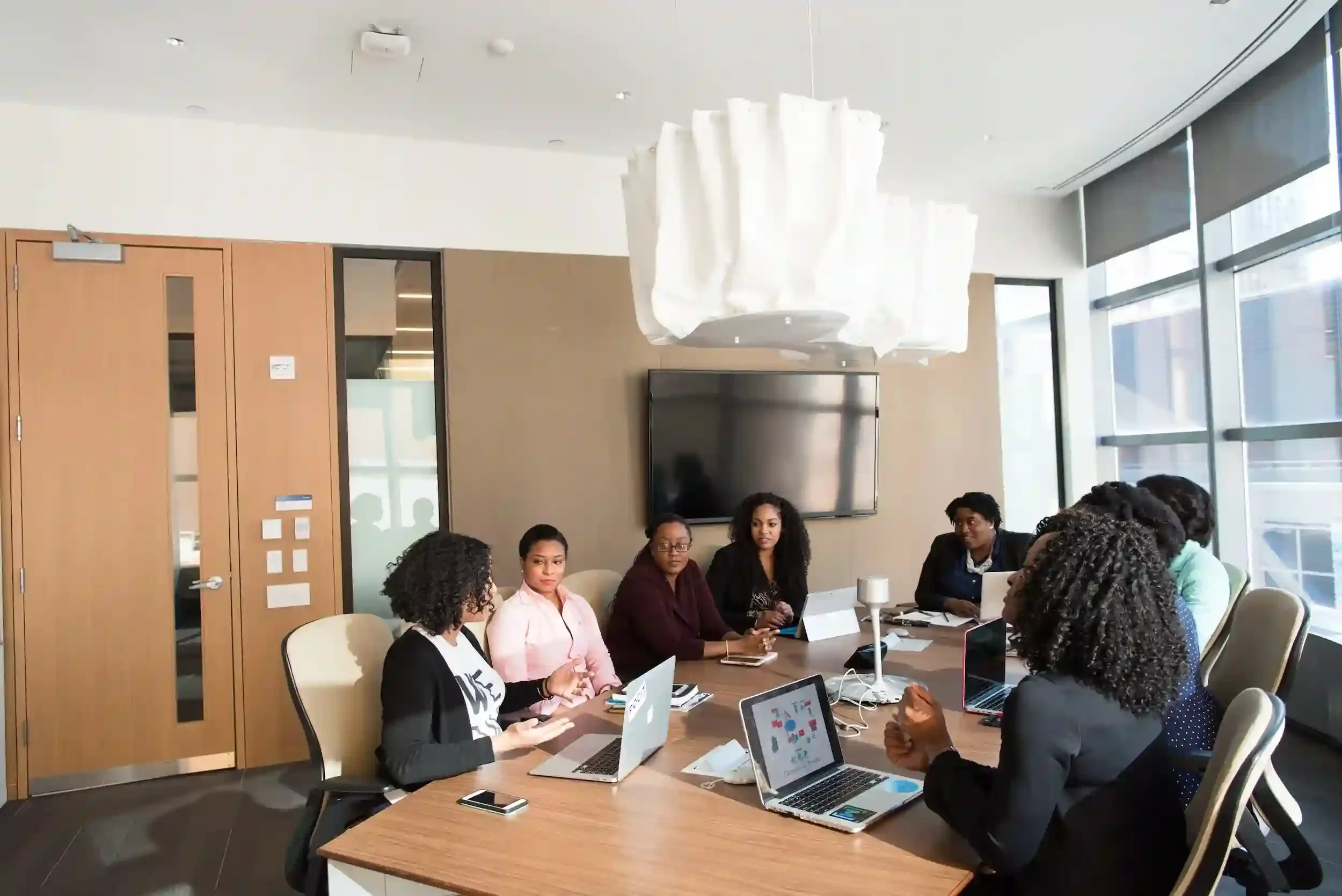 A diverse group of women sitting around a conference table in a modern office, engaged in a meeting or discussion, with laptops and tablets in front of them, large windows and a television screen on the wall behind.