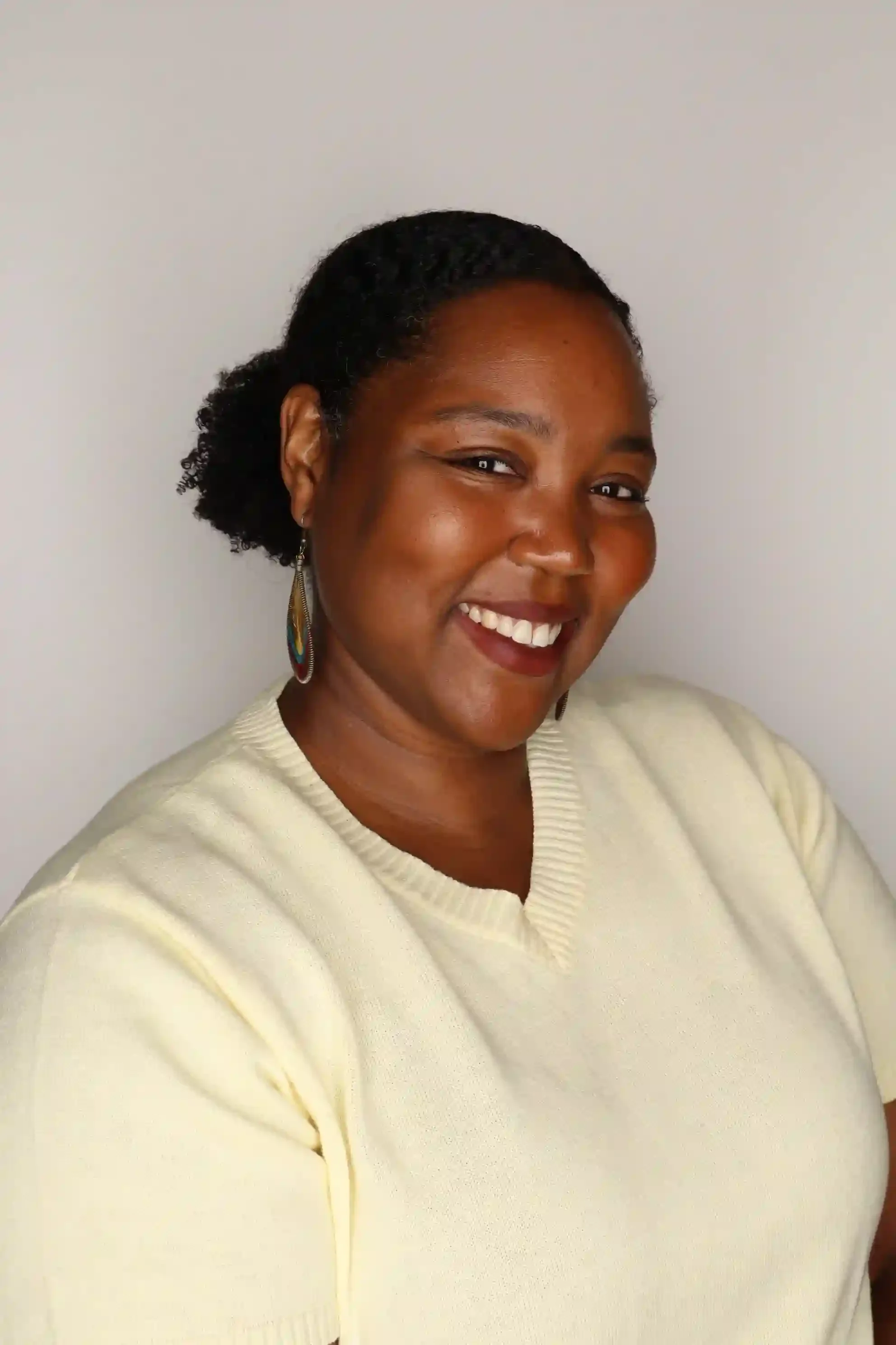 A woman smiling with her hair styled in short natural curls, wearing a cream-colored sweater and feather earrings with blue, green, and yellow accents, standing against a plain light-colored wall.