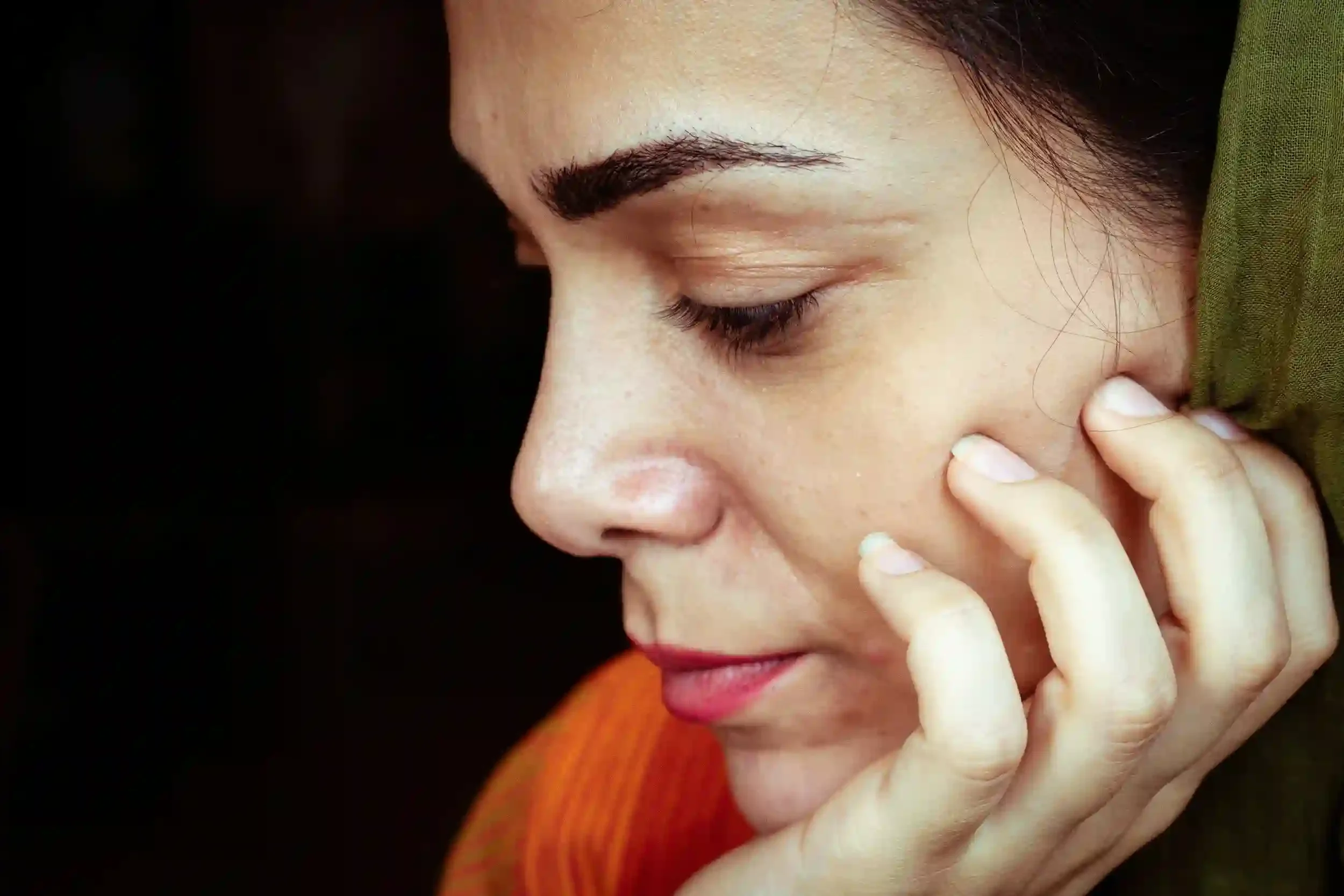 Close-up of a woman with dark hair, eyes closed, holding her face with her fingers, showing her eyebrows, eyelashes, and part of her nose and lips.