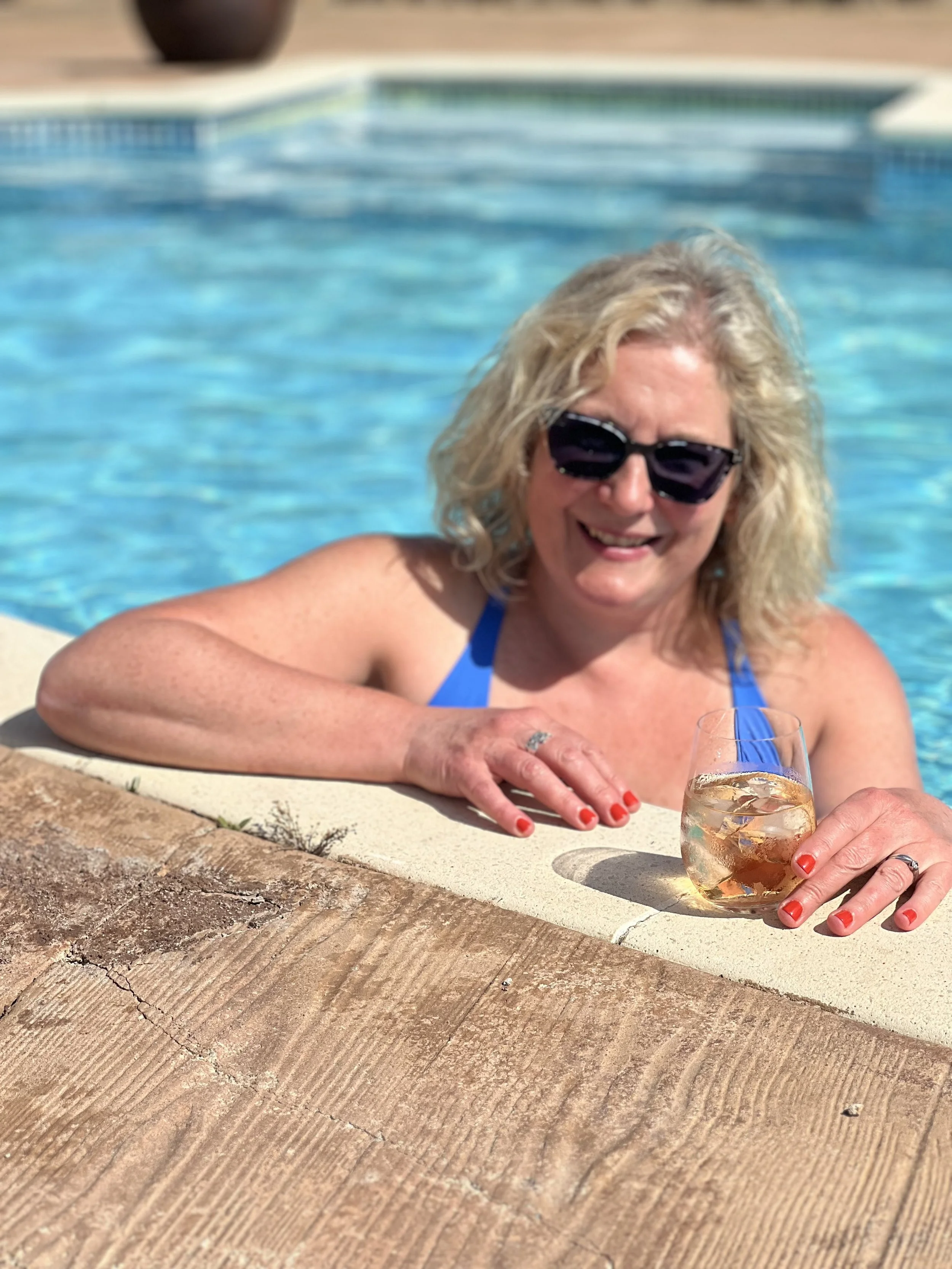 A smiling blonde woman wearing sunglasses relaxes in a turquoise swimming pool while holding a stemless glass of rosé wine served over ice cubes.