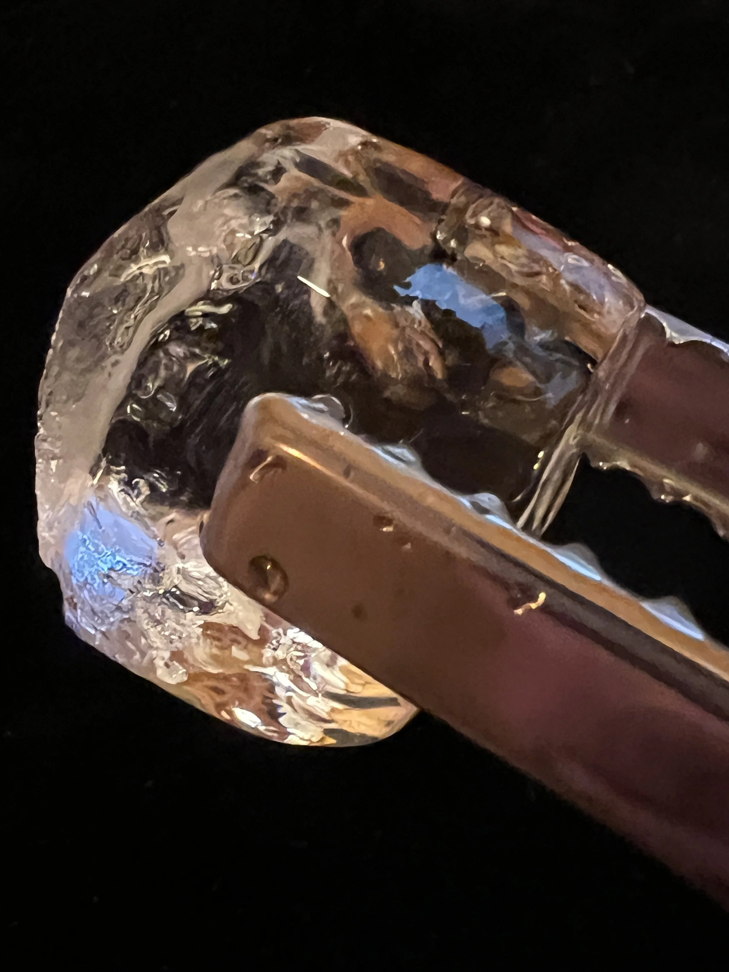 A close-up macro shot of shiny metallic tongs picking up a large, crystal-clear, dense ice cube against a dark background.