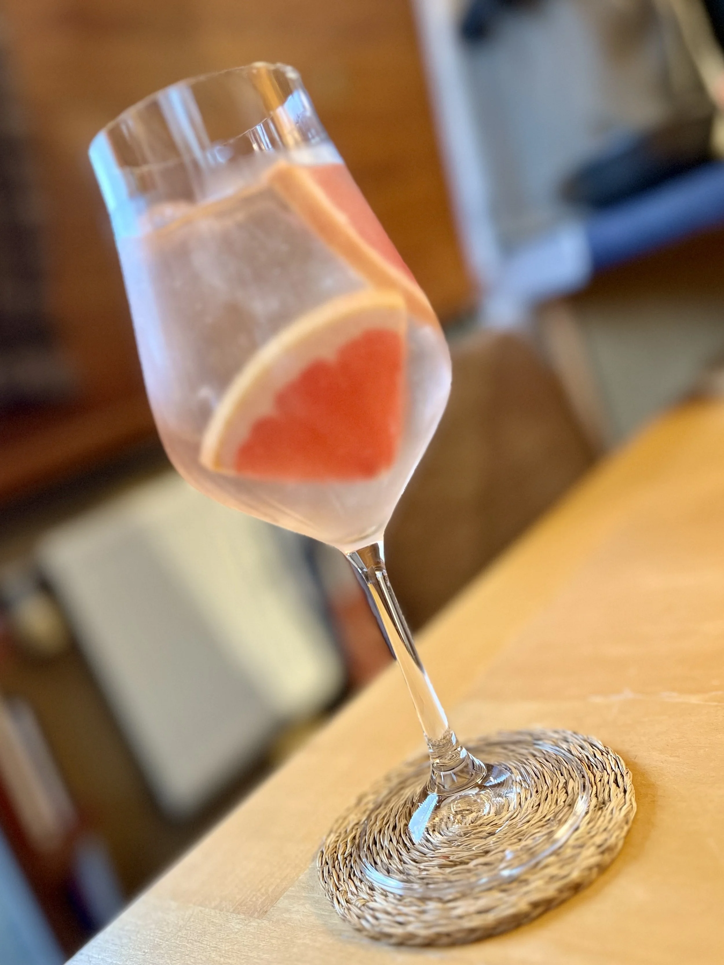A tall, elegant wine glass filled with a chilled, pale pink rosé wine, ice cubes, and a fresh slice of pink grapefruit garnish, sitting on a woven coaster.