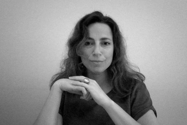 Black and white photo of a woman with long wavy hair, resting her chin on her hand, looking at the camera with a neutral expression.