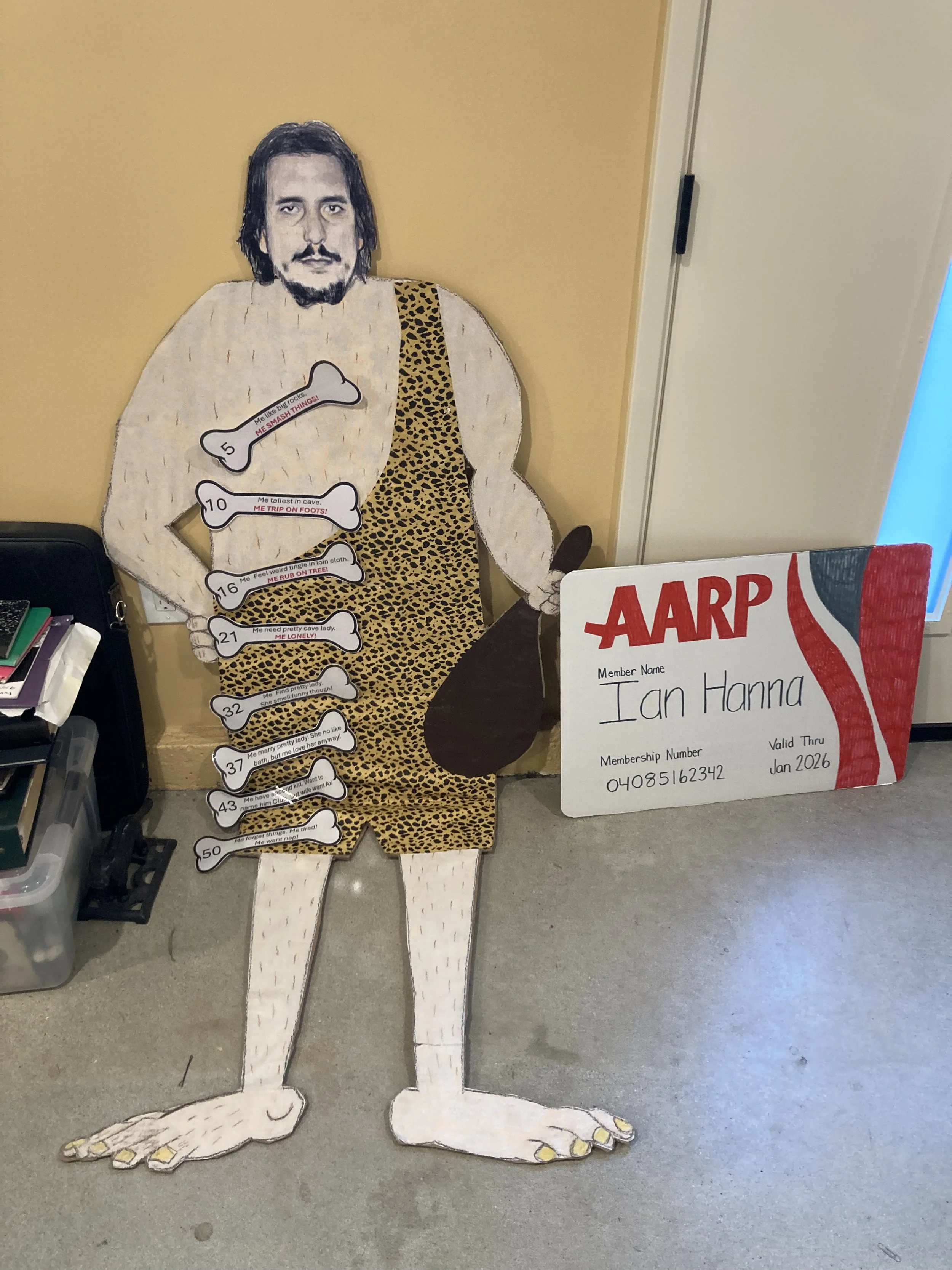 A large, cartoon-style cutout of a person with long black hair, a mustache, and beard, wearing a leopard print dress. The cutout is decorated with several bones that have humorous slogans written on them. There is a signboard with the name 'Ian Hanna