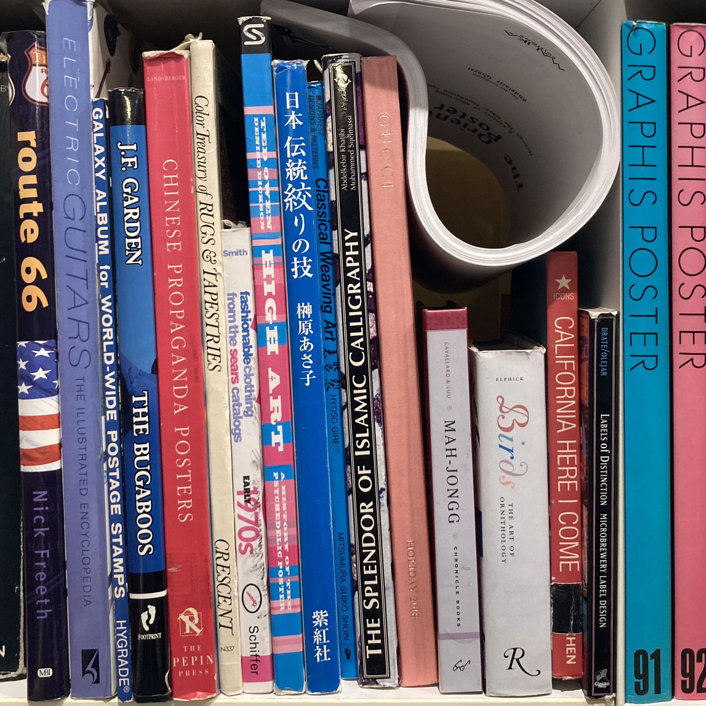 Shelf with a variety of books, some with visible titles including 'Gala', 'Joe', 'The Department of Peps', and 'Graffiti'. Also visible is a roll of white paper or poster, some boxes, and miscellaneous items.