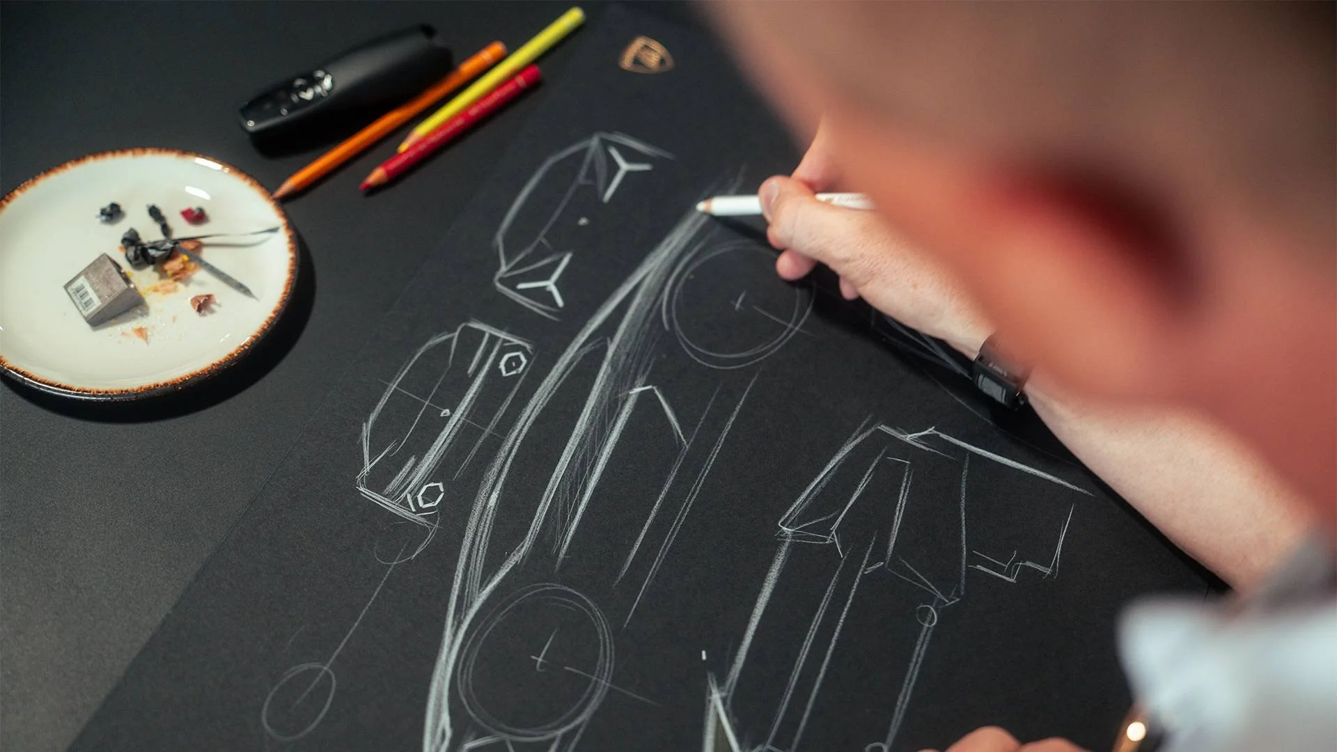 Person drawing a technical design of a vehicle on black paper with white chalk or pastel, with various colored pens and a small plate holding chalk pieces and scraps nearby.