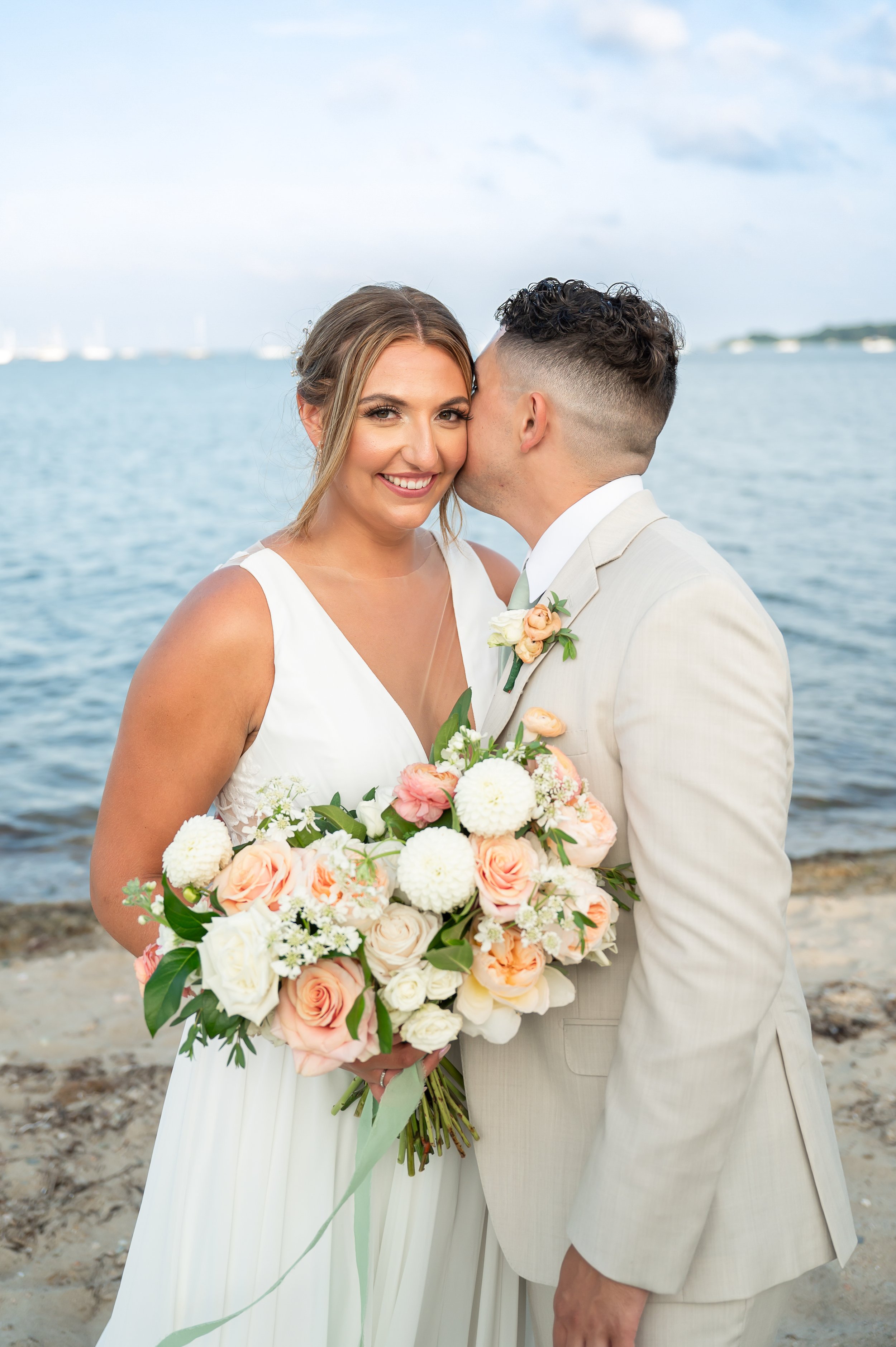A bride and groom on the beach, with the groom kissing the bride's cheek. The bride holds a large bouquet of white, peach, and pink flowers. The bride wears a white dress, and the groom wears a light-colored suit. The background features water and a cloudy sky.