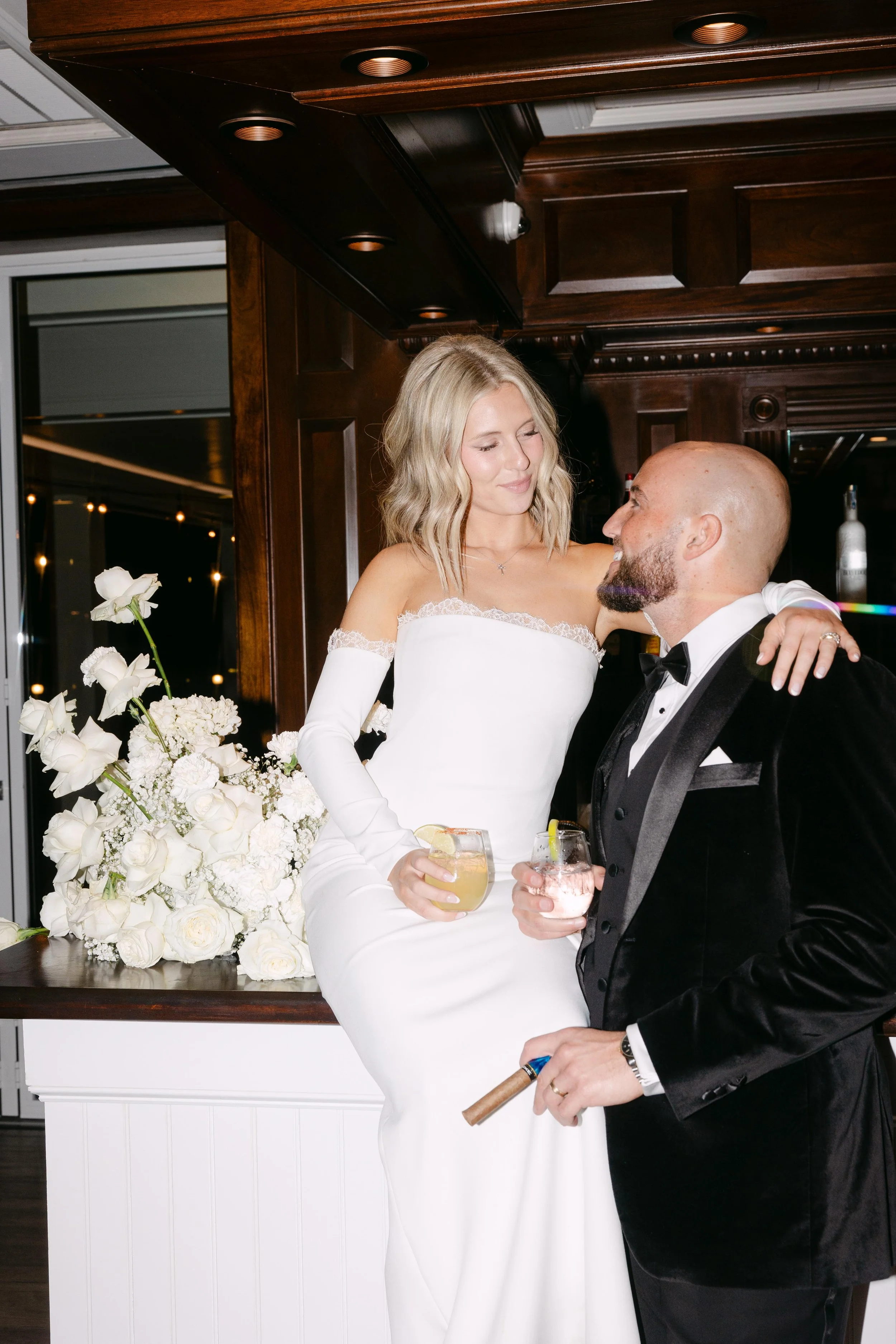 A bride and groom at their wedding, with the bride sitting on a bar counter and the groom standing close, holding a cigar and drinks, in a sophisticated venue with dark wood paneling and floral decorations.