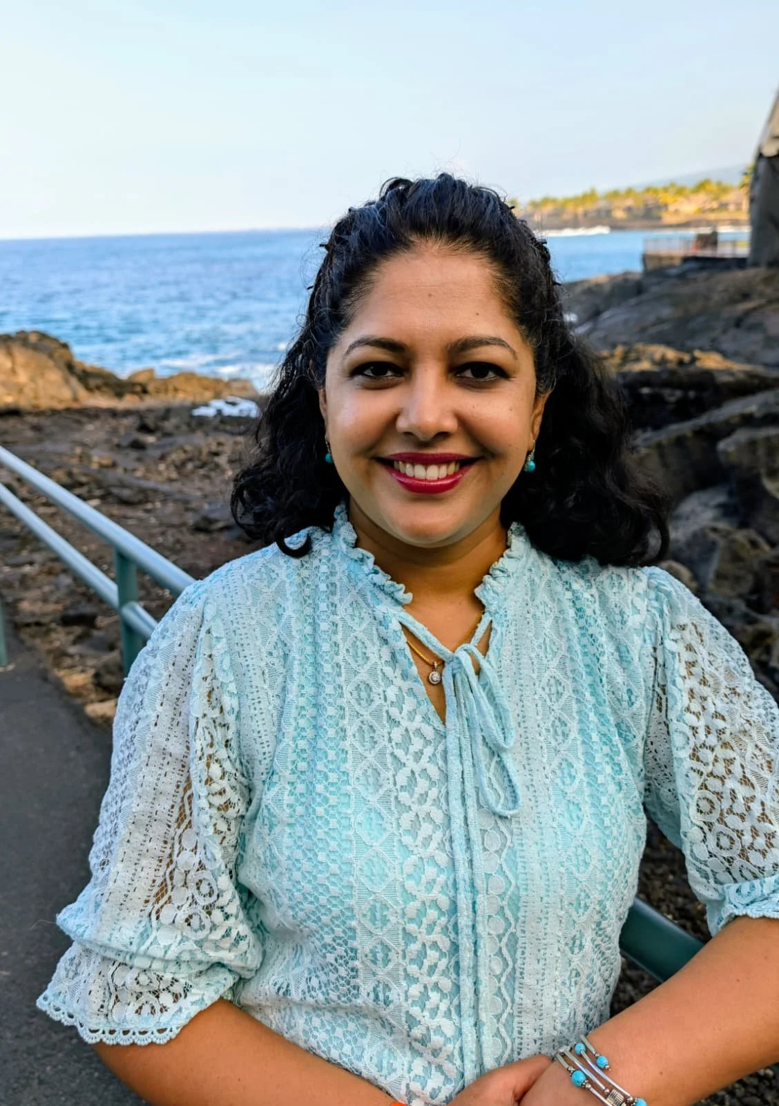 image of a smiling Indian woman with dark brown hair, light brown skin, and light blue shirt. She is standing in front of rocks on a coastline and there is water behind her